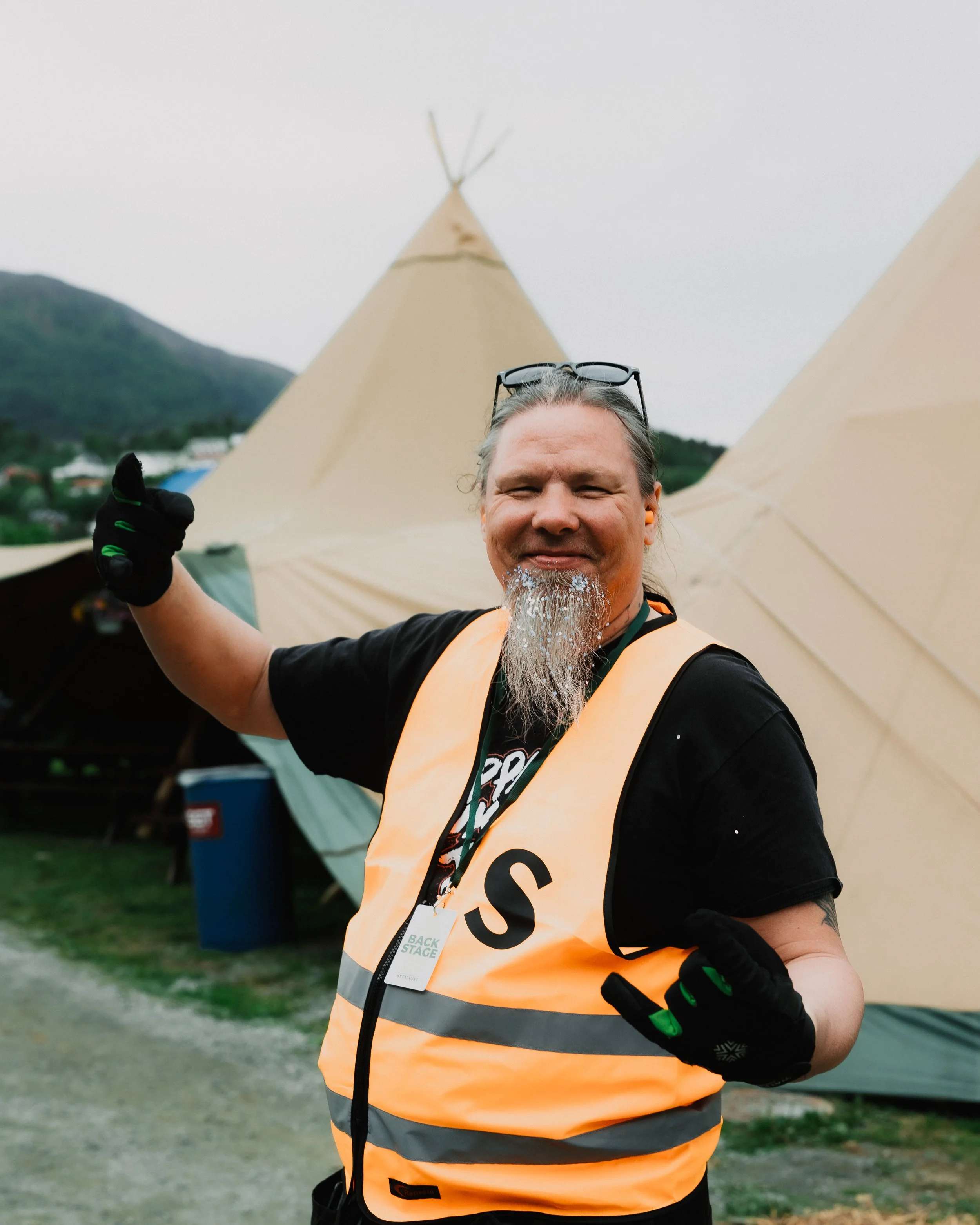 En mann som smiler med vann som drypper fra skjegget, iført refleksvest og hansker, foran et teppe av campingtelt.