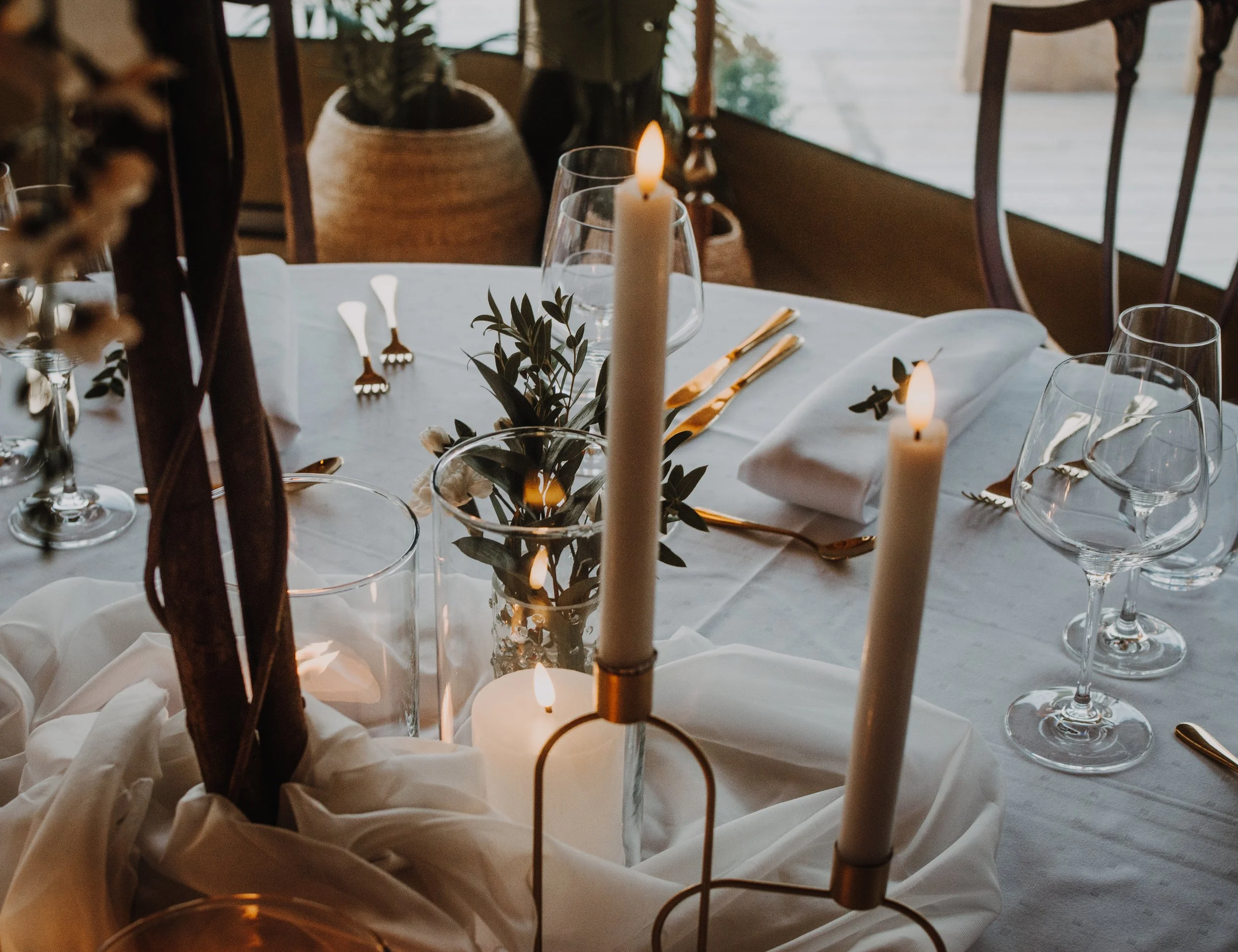 Et bord dekket for et måltid med hvit duk, tallrike vin- og vann glass, gullbestikk, hvite servietter, lysestaker med tente lys og grønne blader som dekorasjon. Rømmelig belysning med naturlig lys som kommer inn gjennom et vindu.