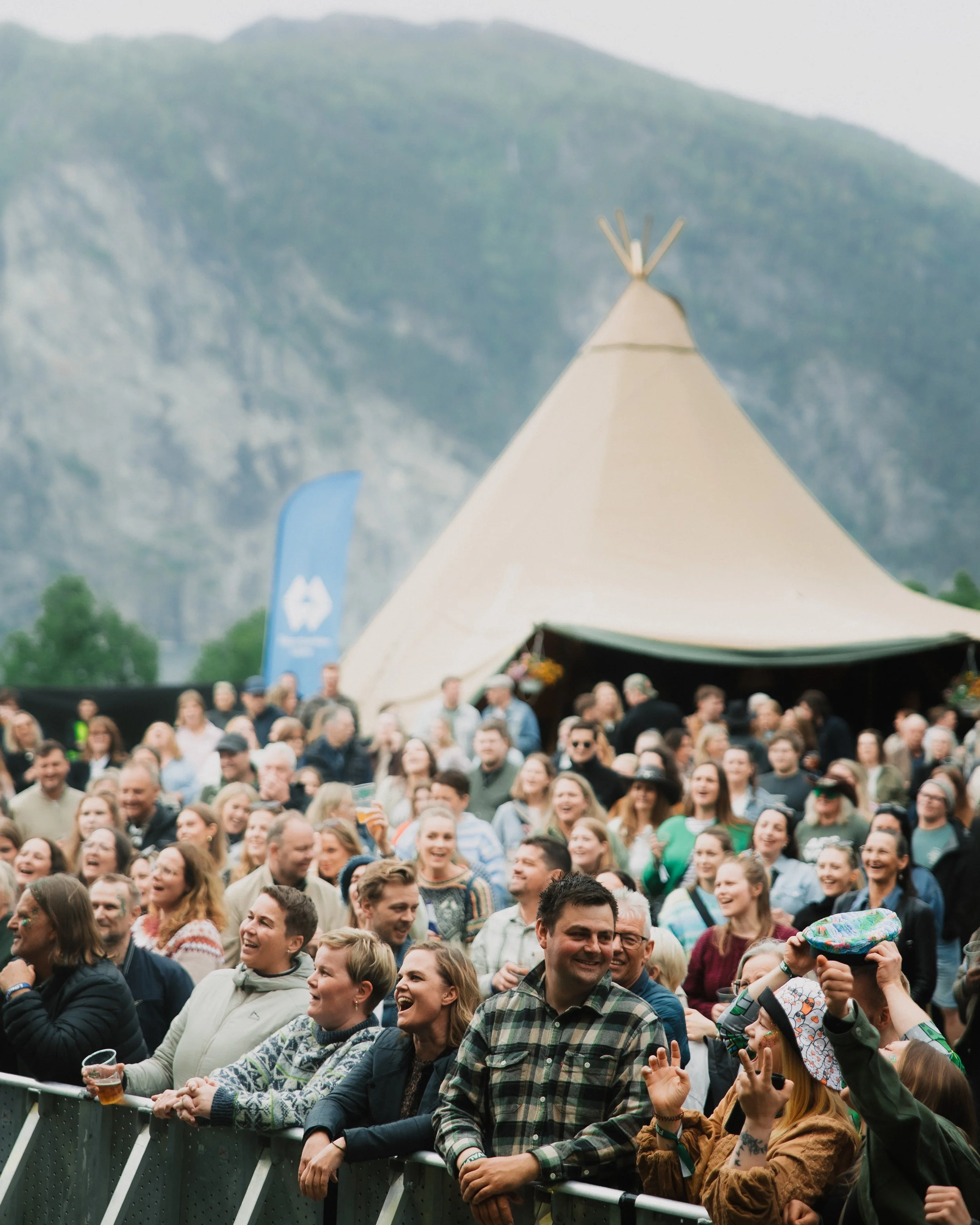 En stor folkemengde foran en scene, de ler og koser seg, i bakgrunnen er fjell og en stor teepee. Det ser ut til å være en utendørs festival eller konsert.