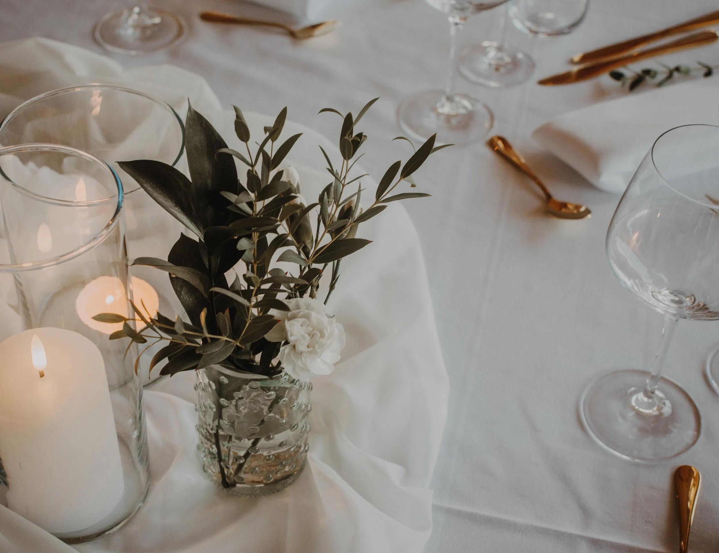 Dekket bord med glass, sølvskjeer, små lys i glasslykter, en liten blomsterbukett med grønne blader i en krystallvase, og hvitt serviett.