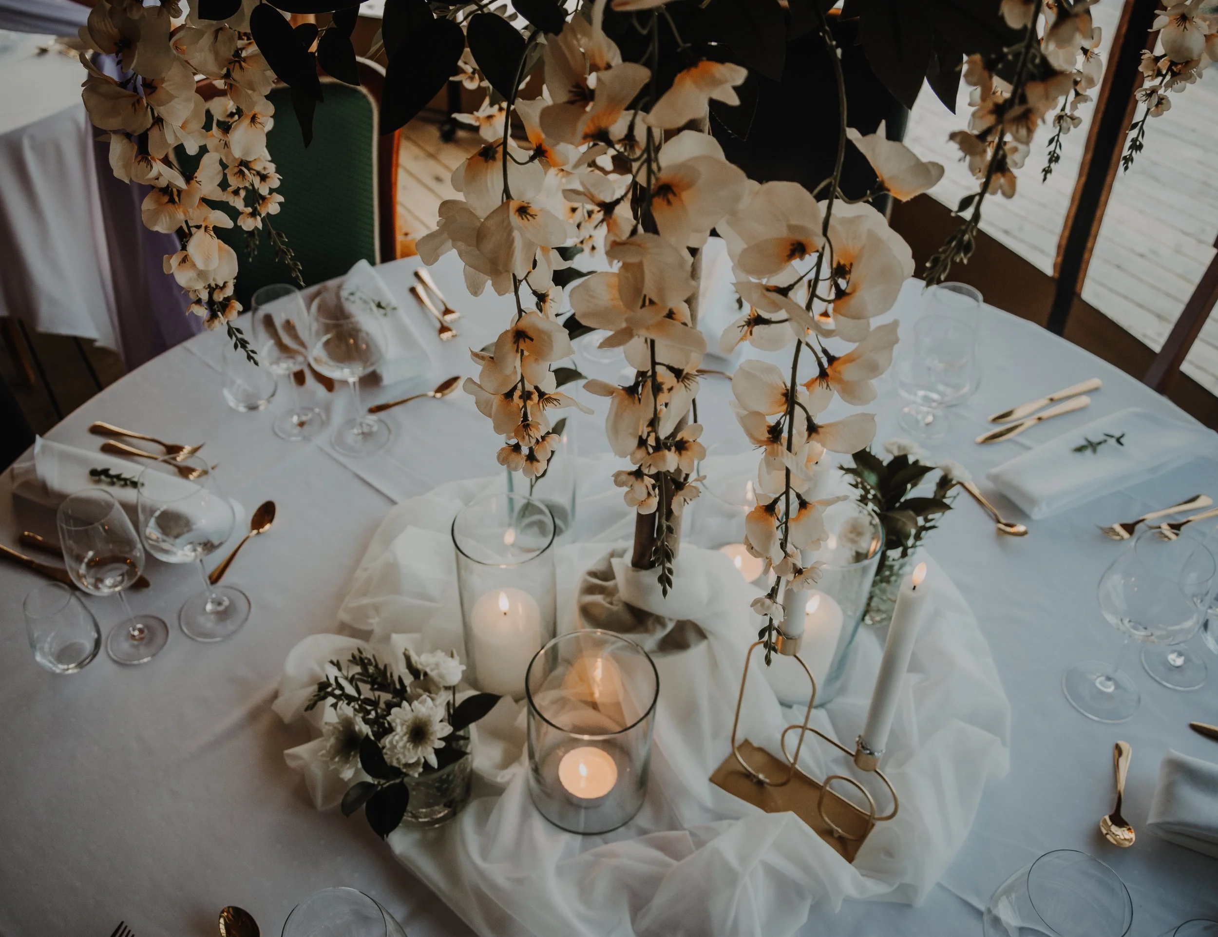 Et dekket bord med hvit duk, glass, sølvbestikk og blomsterdekorasjoner, inkludert hvite blomster og lys, under en takketg i et innendørs rom.