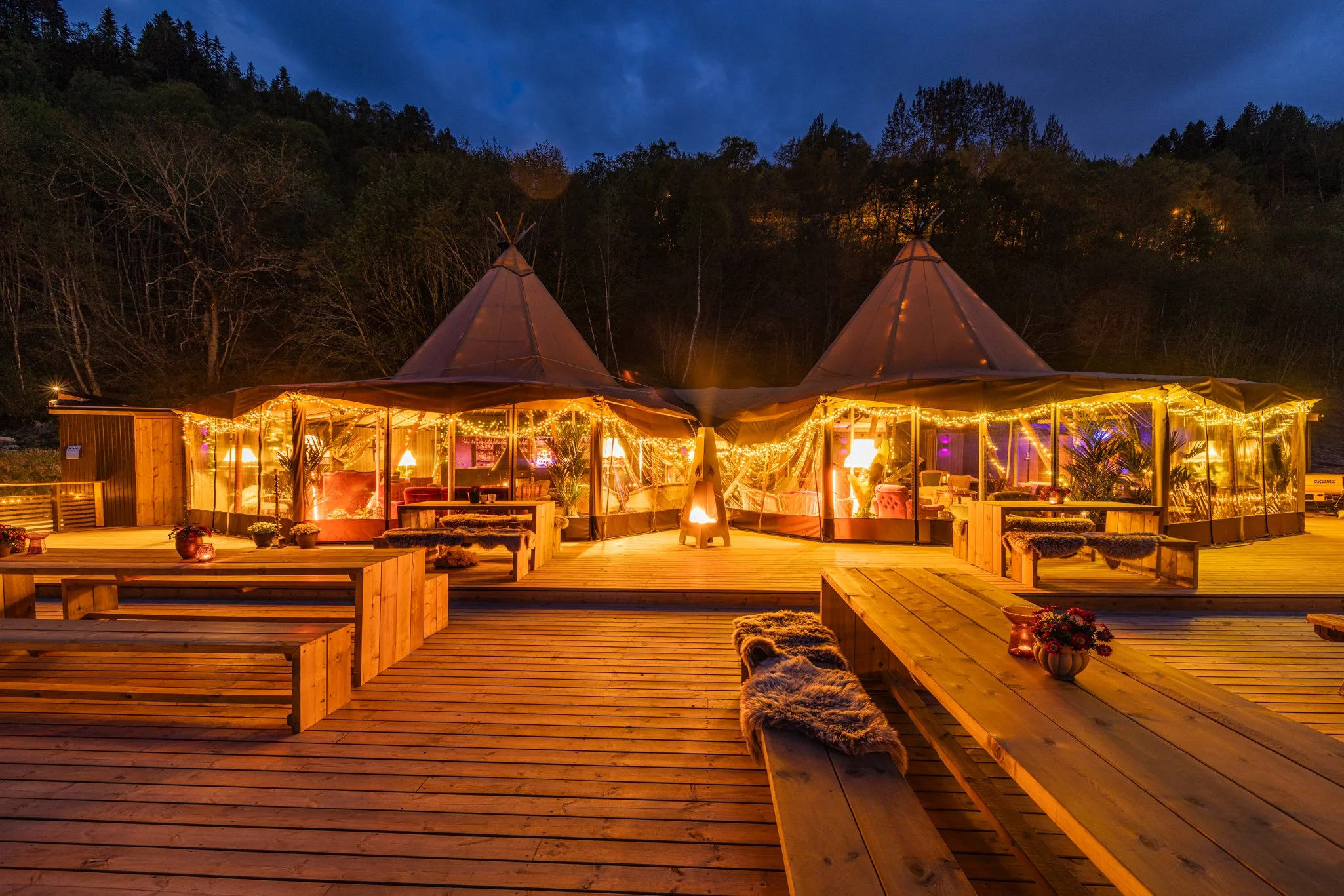 Utsiden av en lyssatt bohemisk tipi-overnattingsplass ved skogkledde fjell, med tre tipi-telt og sitteplasser på en treterrasse om kvelden.
