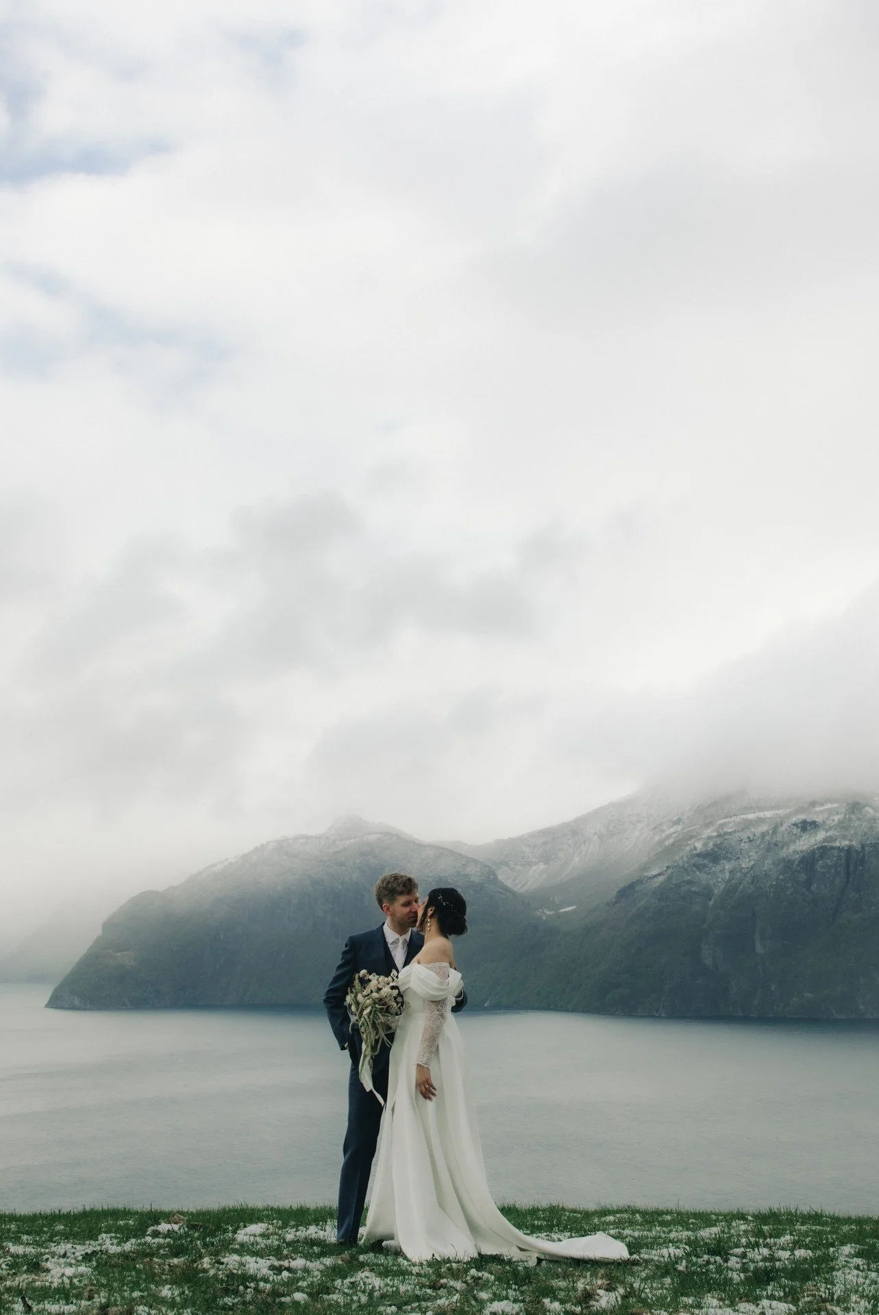 Et brudepar står ved en innsjø omgitt av fjell, under en overskyet himmel, og ser på hverandre