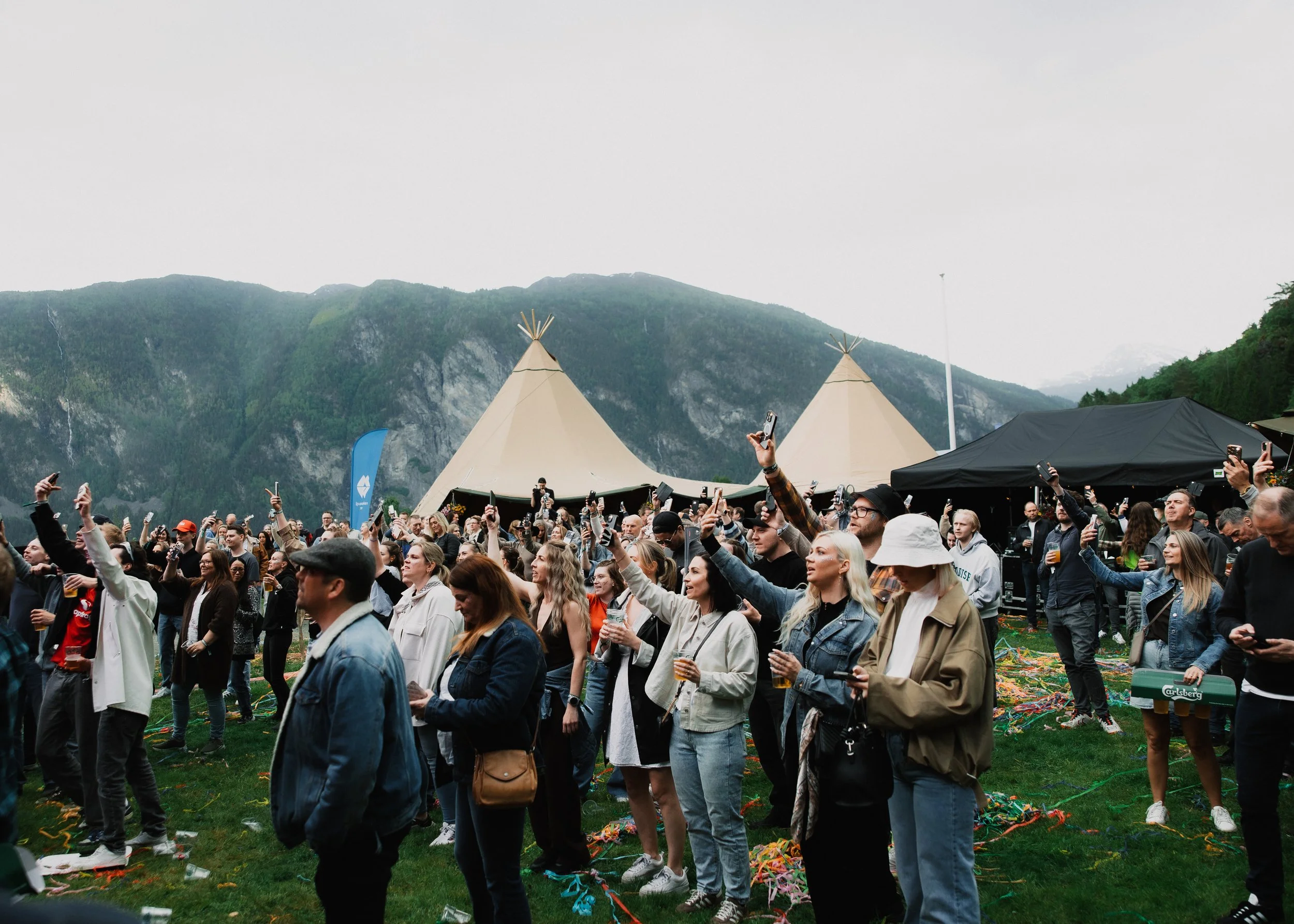 En stor utendørs konsert med mange mennesker som danser og tar bilder mot en bakgrunn av fjell og telt.