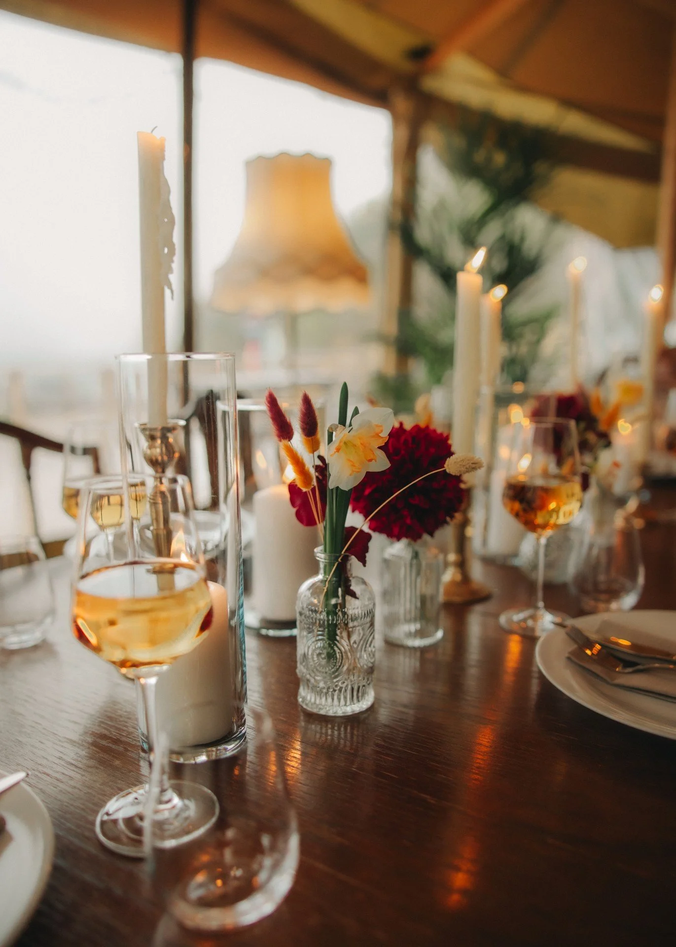 Et bord dekket for fest eller middag med blomsterarrangementer, stearinlys og glass med vin, i en koselig innendørs setting.