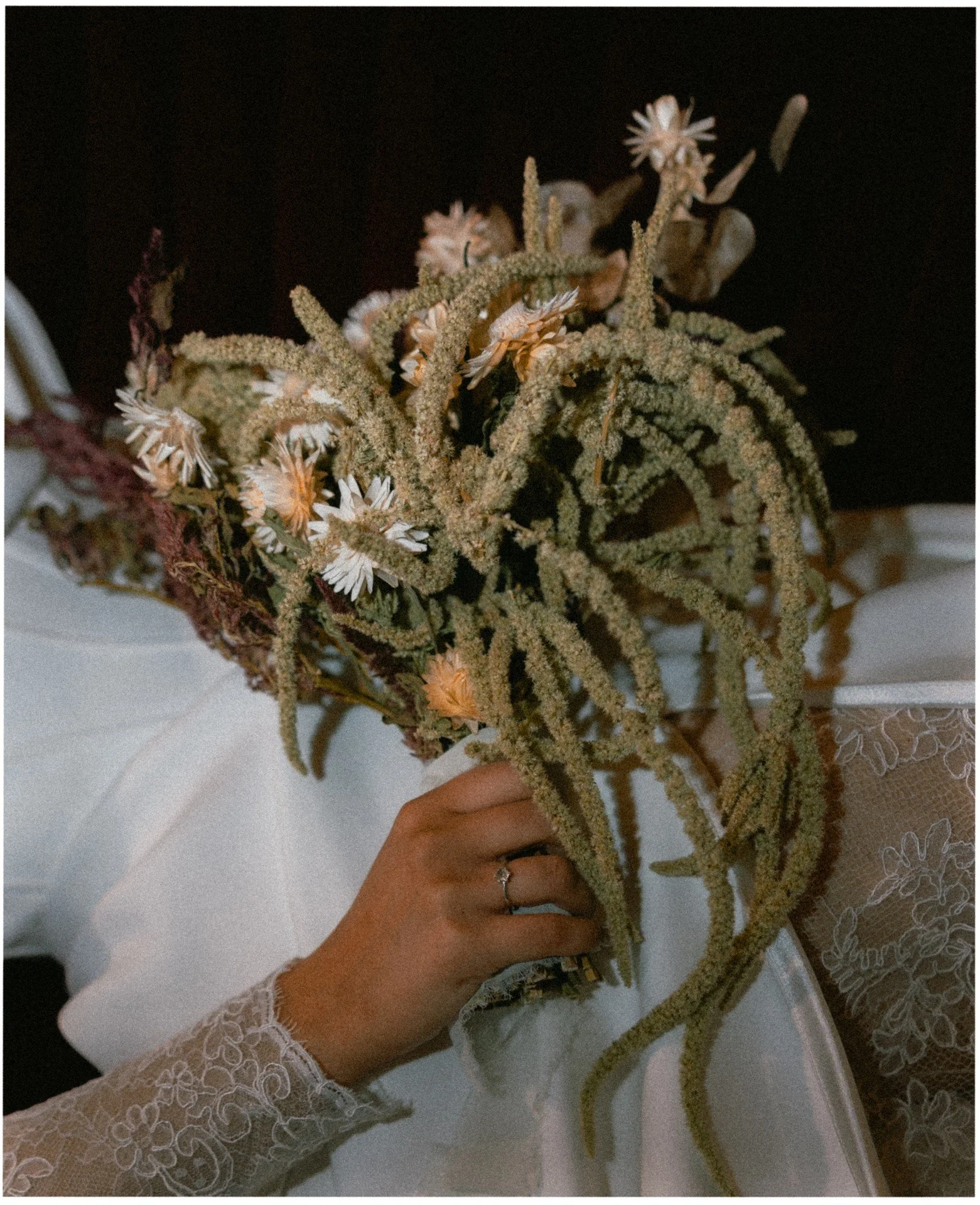 En person holder et brudebukett med tørkede blomster og planter, inkludert kornsløyfer og keramiske blomster, mot en hvit bordduk med blomstermønster.