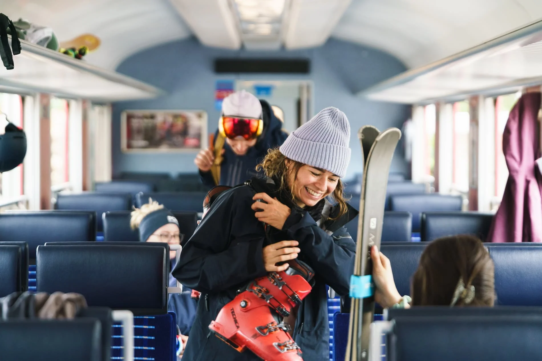 Menschen in einem Zug, zuletzt eine lachende Frau, die Skischuhe hält und sich mit anderen unterhält, während sie Ski trägt.