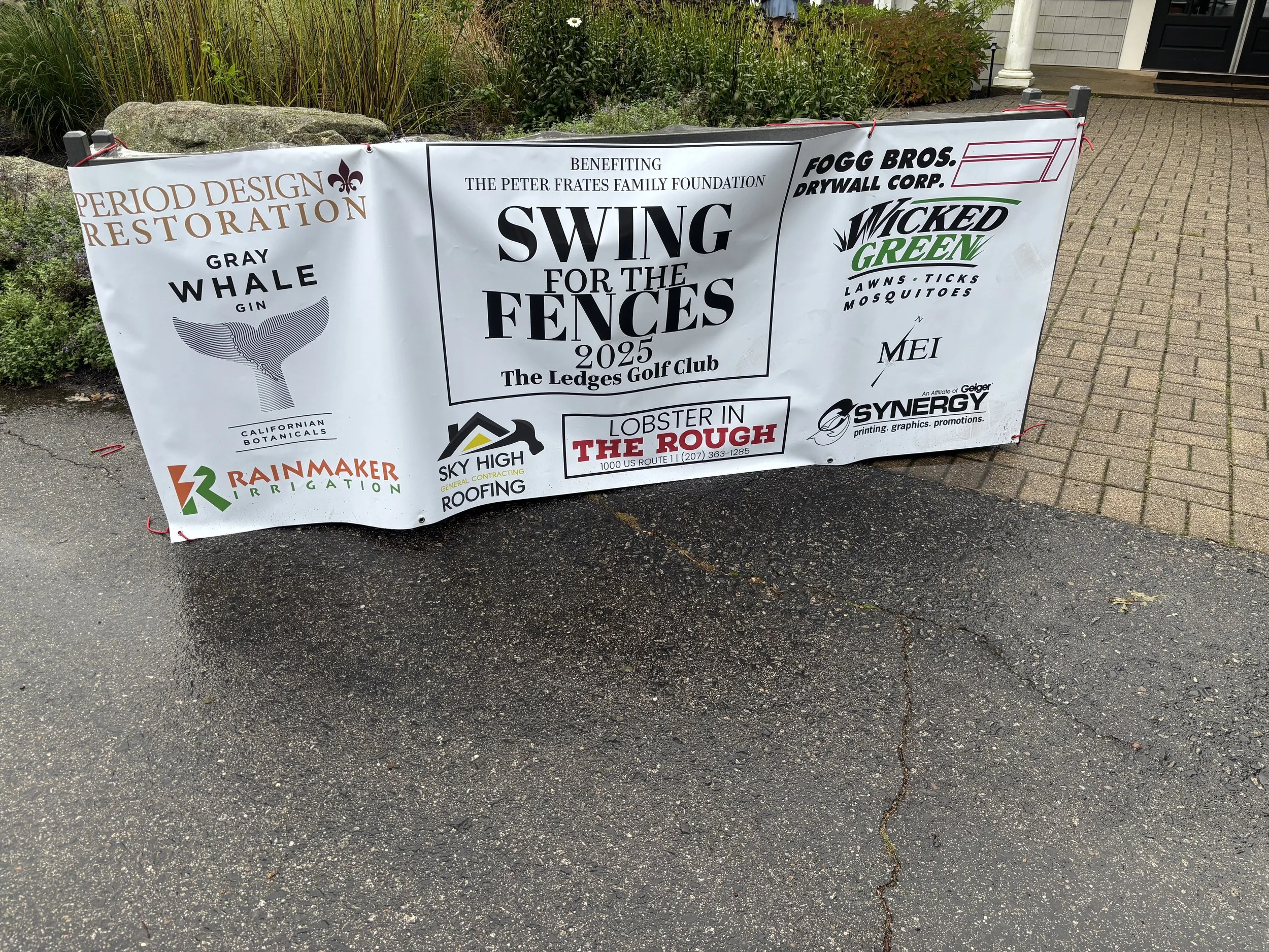 A large white advertisement banner secured with red ties, placed on a paved walkway in front of a flower bed and near a house entrance. The banner promotes a swing event benefitting the Peter Frates Family Foundation, with various sponsor logos and event details.