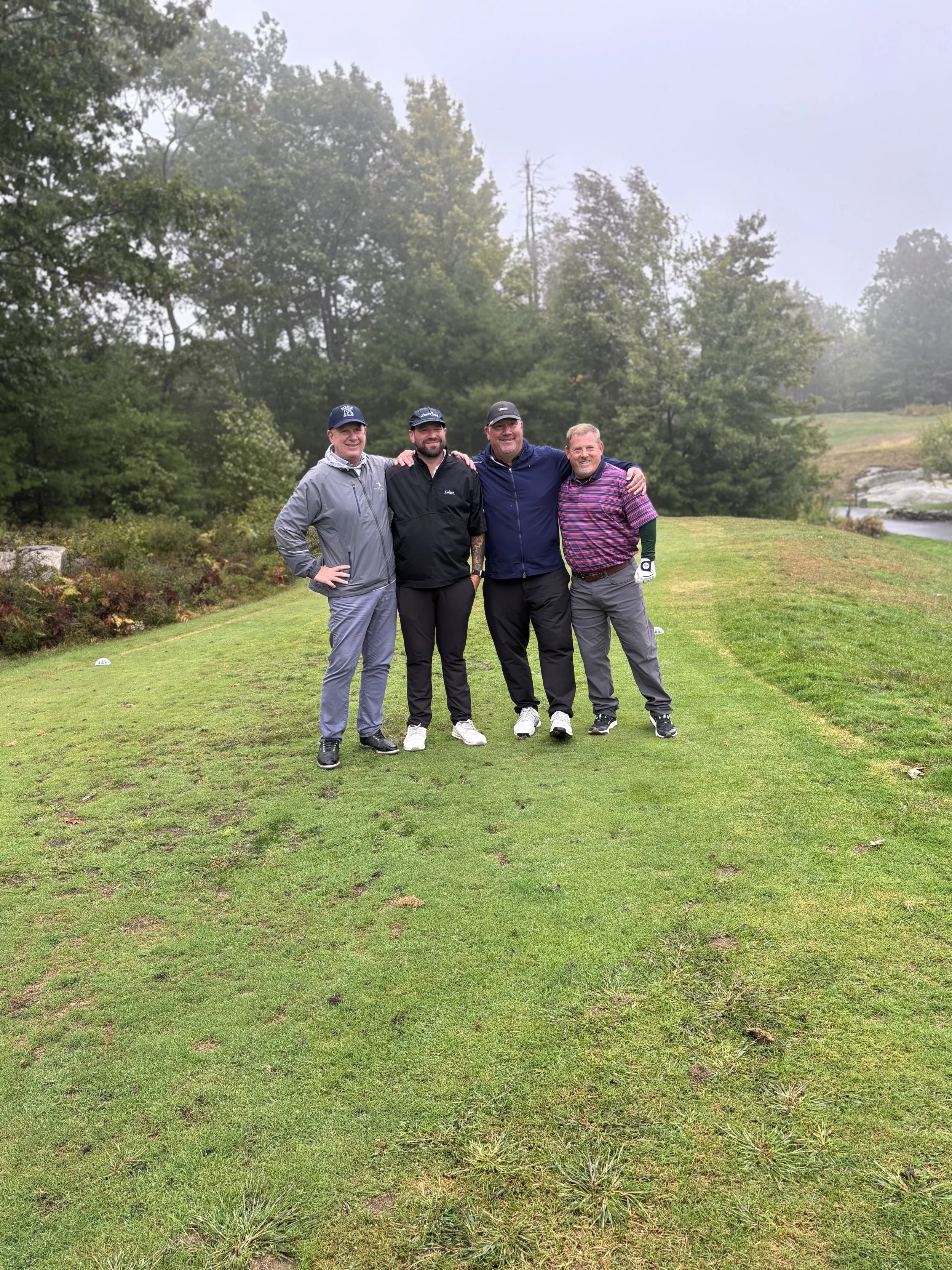 Four men standing on a golf course, smiling, with trees in the background and a misty sky.