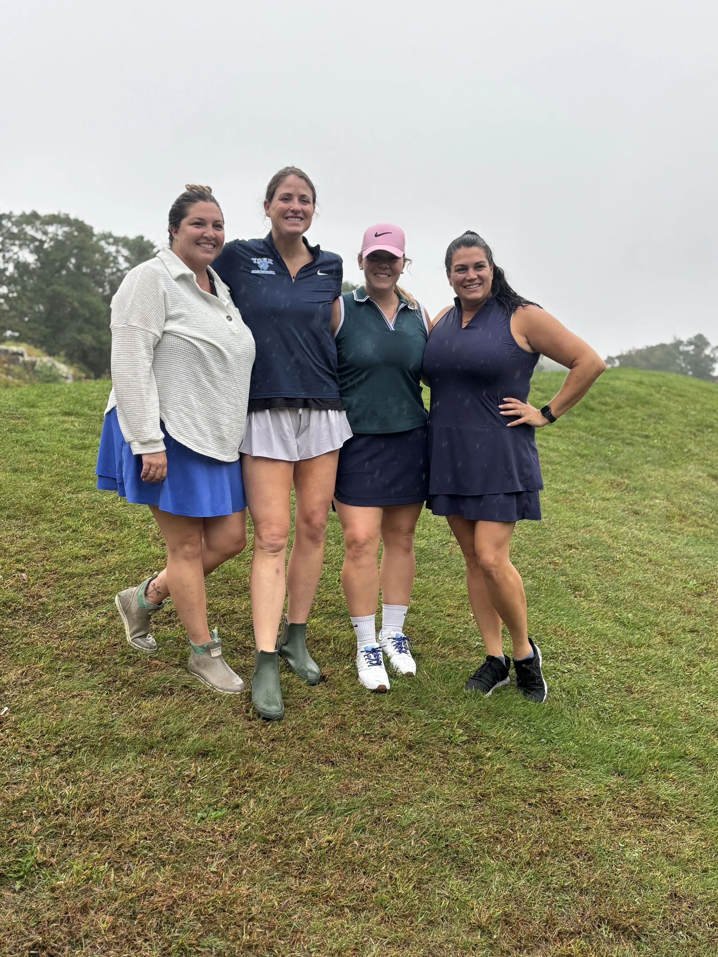 Four women standing on a grassy field, dressed in sportswear, smiling at the camera, in an outdoor setting with trees and cloudy sky in the background.