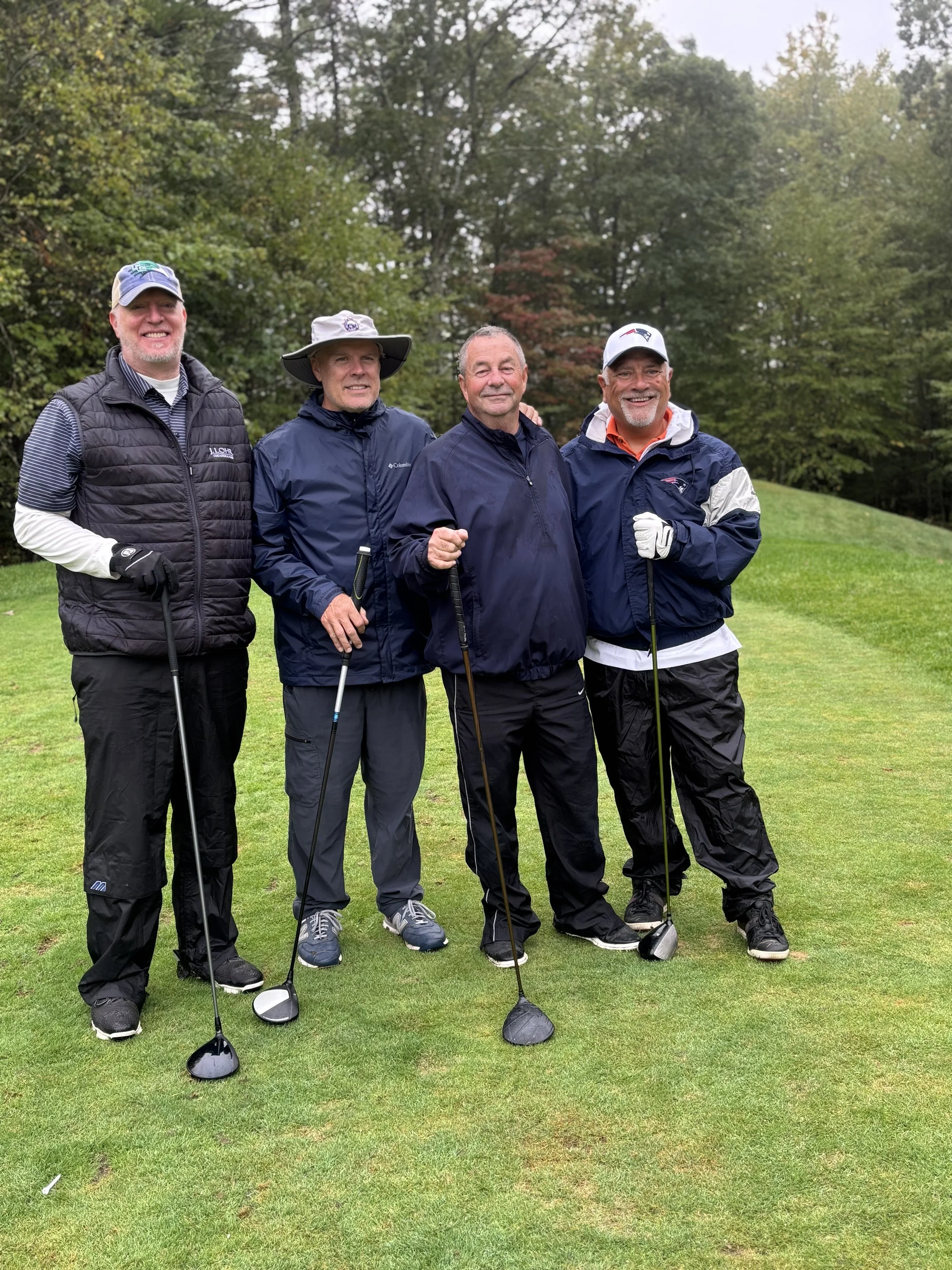 Four men standing on a golf course, holding golf clubs, dressed in golf attire, with trees and grass in the background during overcast weather.