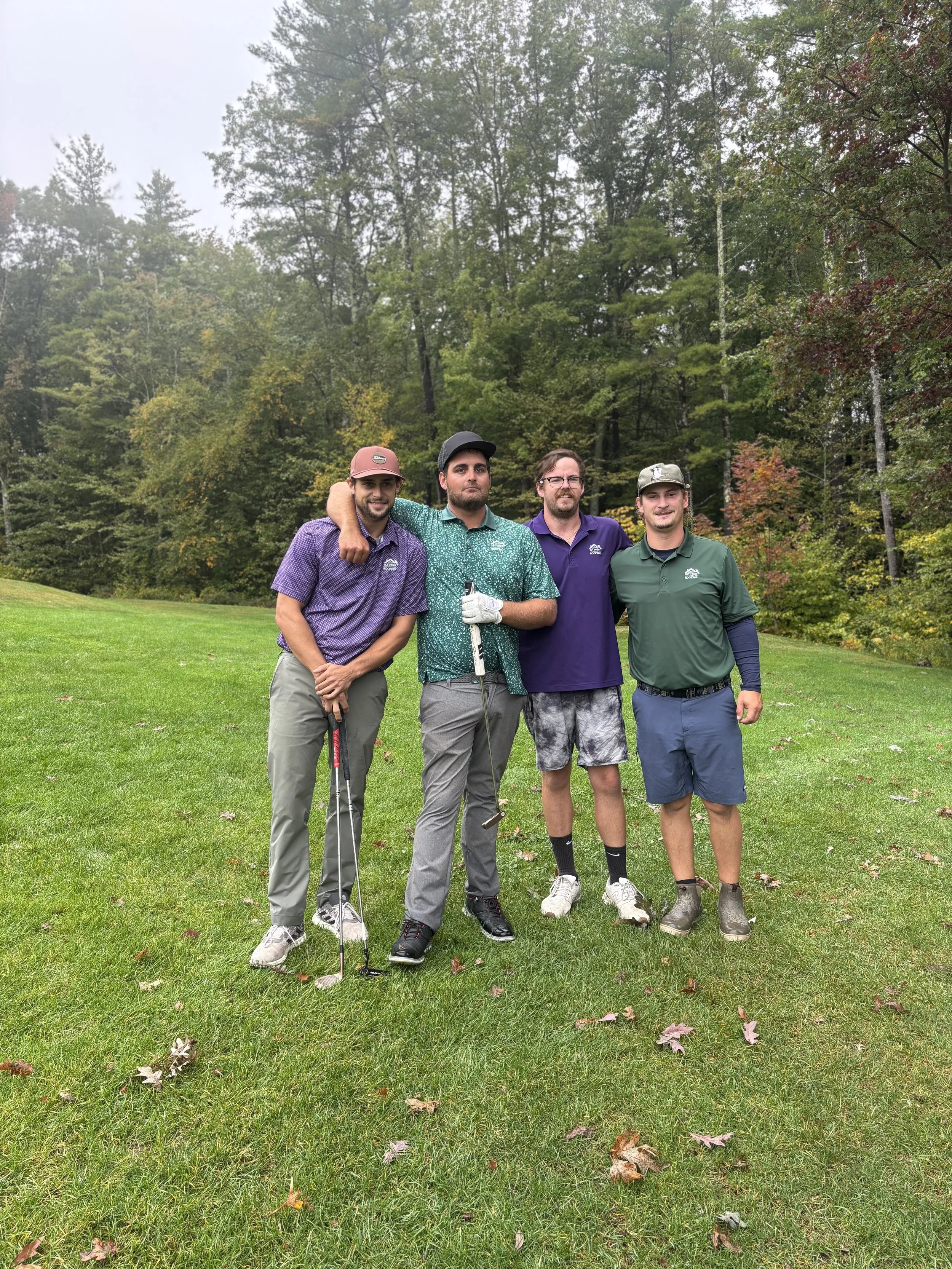 Four men standing on a golf course with trees in the background, dressed in casual golf attire, some holding golf clubs, one wearing a cap, and one with glasses.