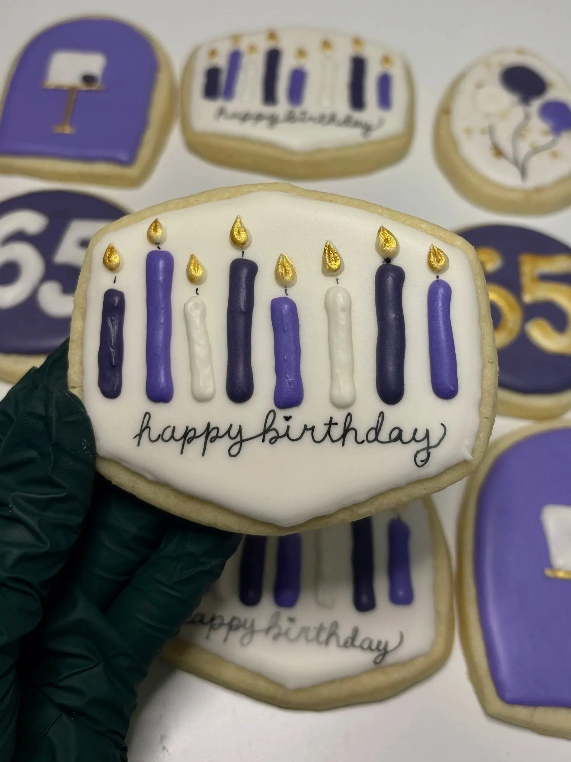 Decorated birthday cookies with purple, white, and gold icing, featuring candles and festive designs, with one cookie in focus that says "happy birthday".