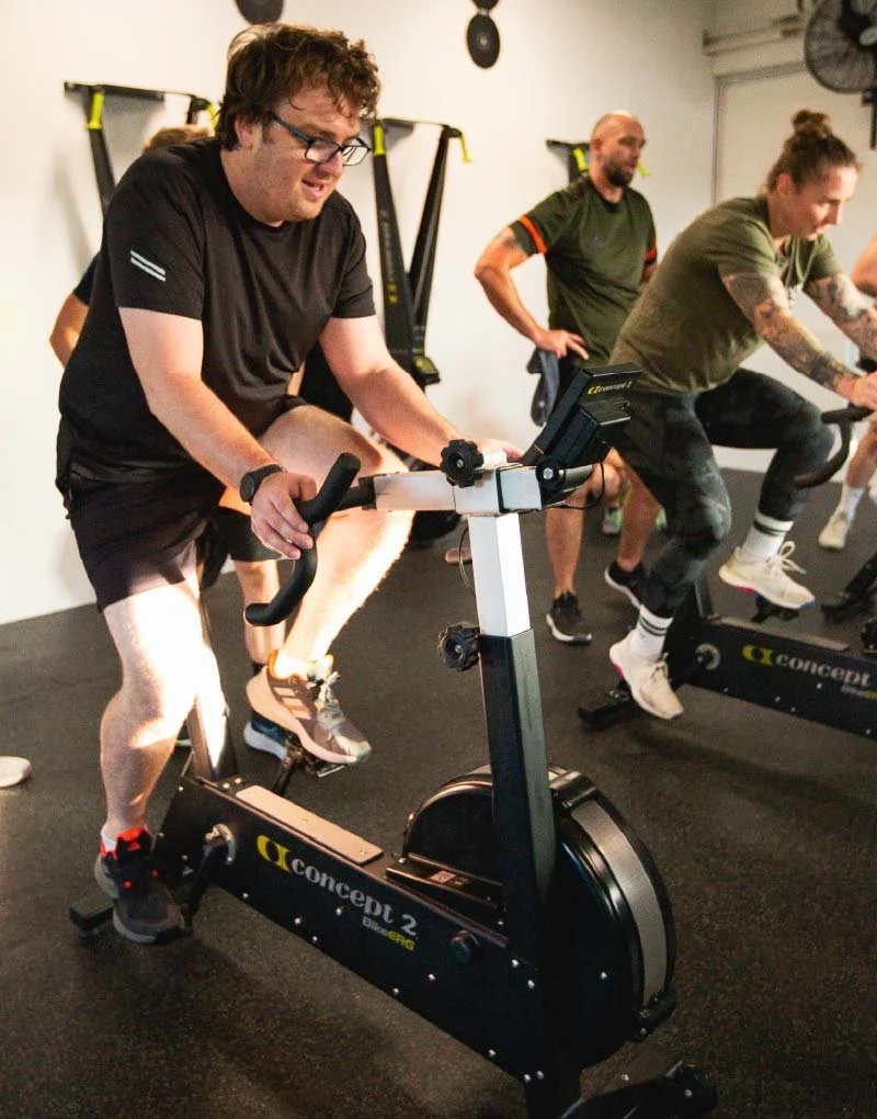 BUILT Training member smiling whilst exercising on stationary bikes in a gym. One man is pedalling on a spin bike, smiling, with two other individuals also riding bikes behind him.