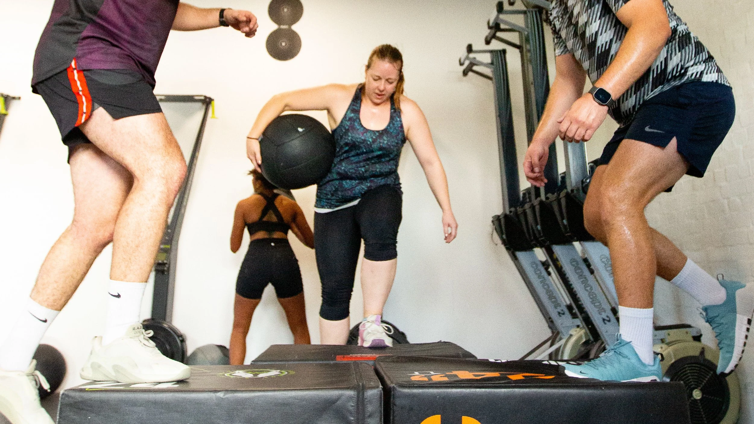 Group of people exercising in a gym, focusing on a woman with a medicine ball, surrounded by others on workout equipment.