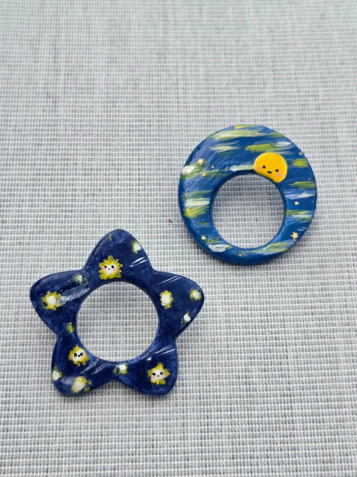 Decorative painted ceramic or plaster rings with cute, cartoonish face designs, one shaped like a flower and the other like a planet with a moon, on a textured light gray surface.