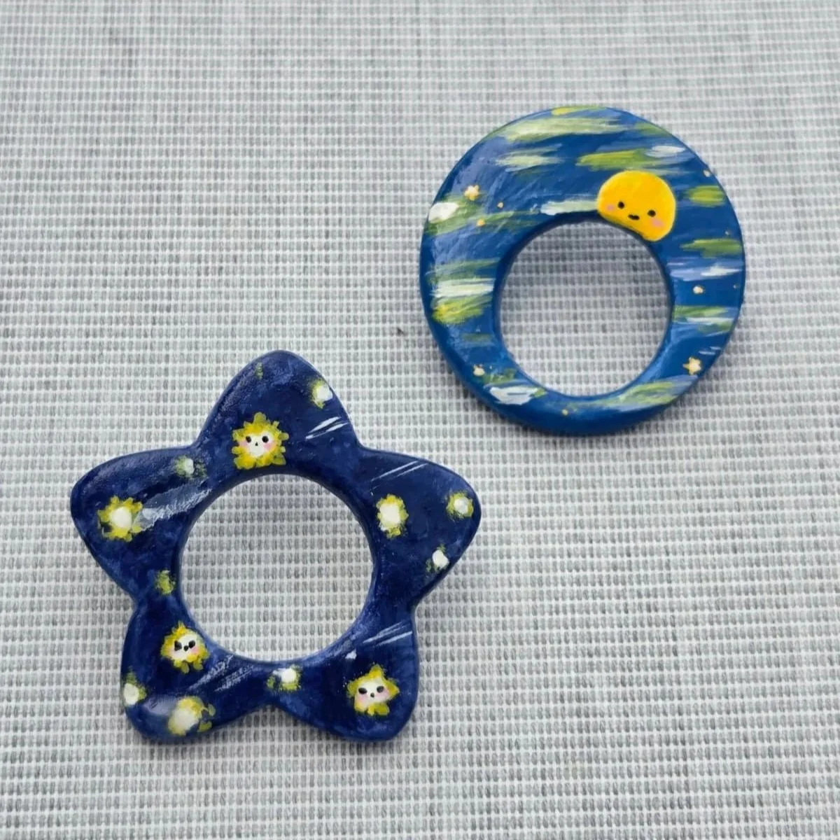 Two decorated ceramic rings with space-themed designs are placed on a gray textile surface. One ring is shaped like a flower and features small smiley faces and yellow accents. The other ring is shaped like a crescent moon and has a smiley face along with clouds and stars painted on it.