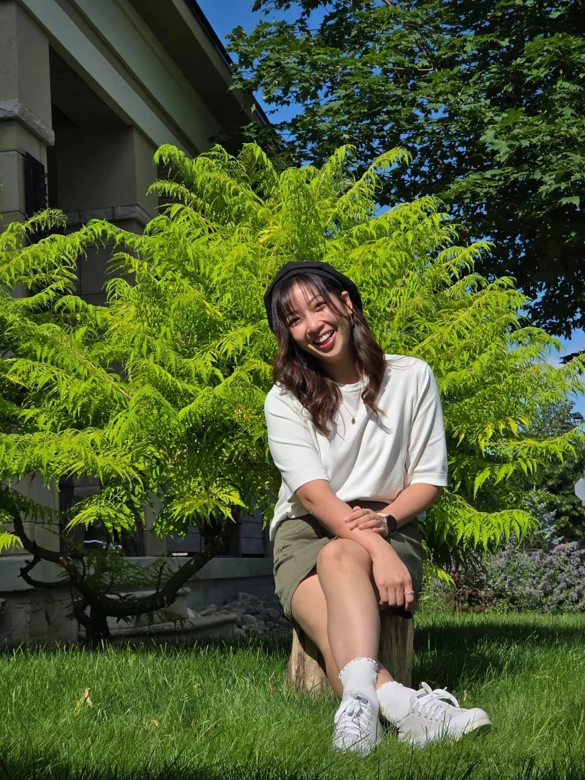 A young woman sitting on a tree stump in front of a lush green bush, smiling and squinting in the sunlight, with a house and clear blue sky in the background.