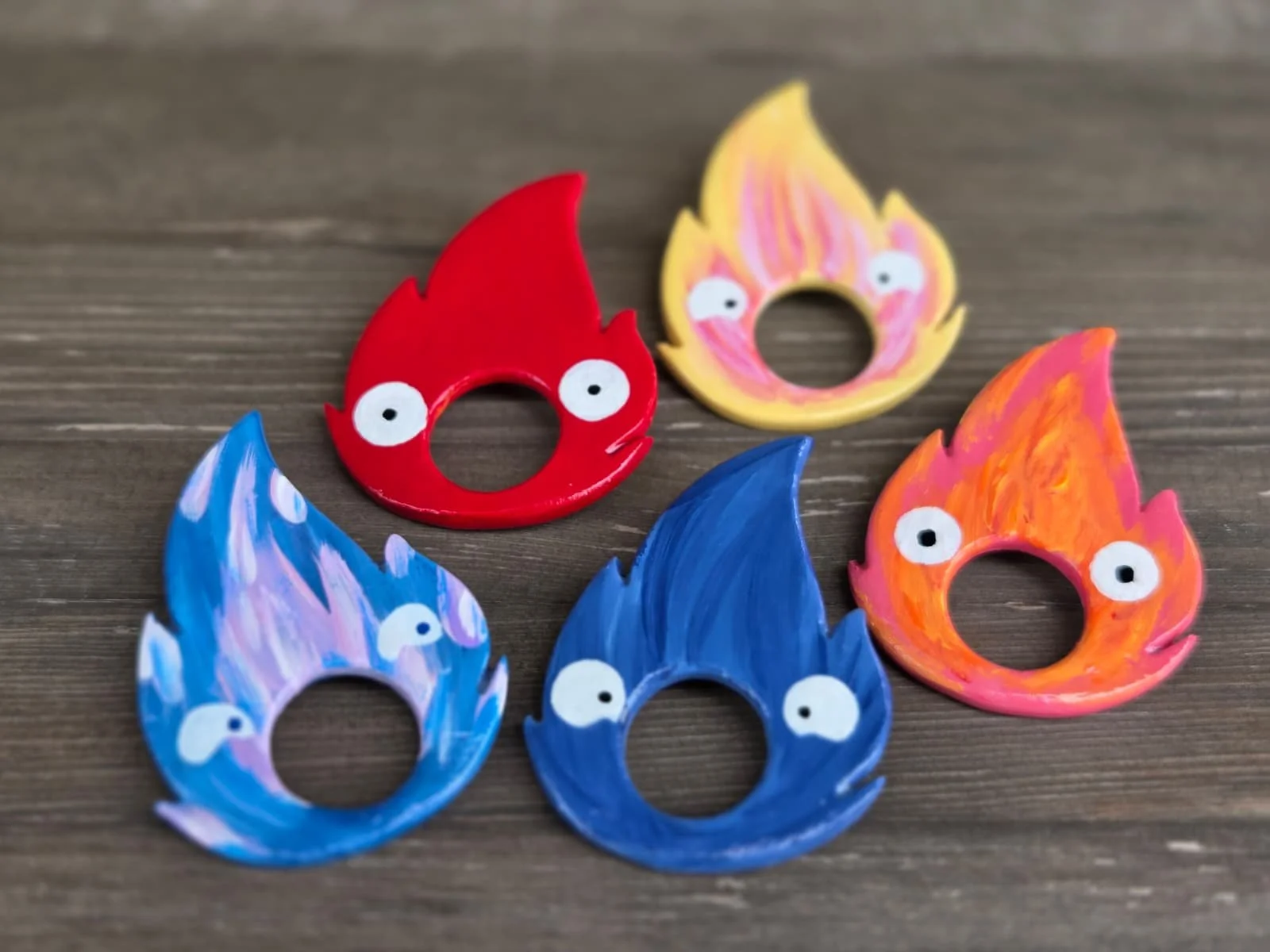 Colored paper flame-shaped cutouts with cartoonish eyes painted on them, arranged on a wooden surface.