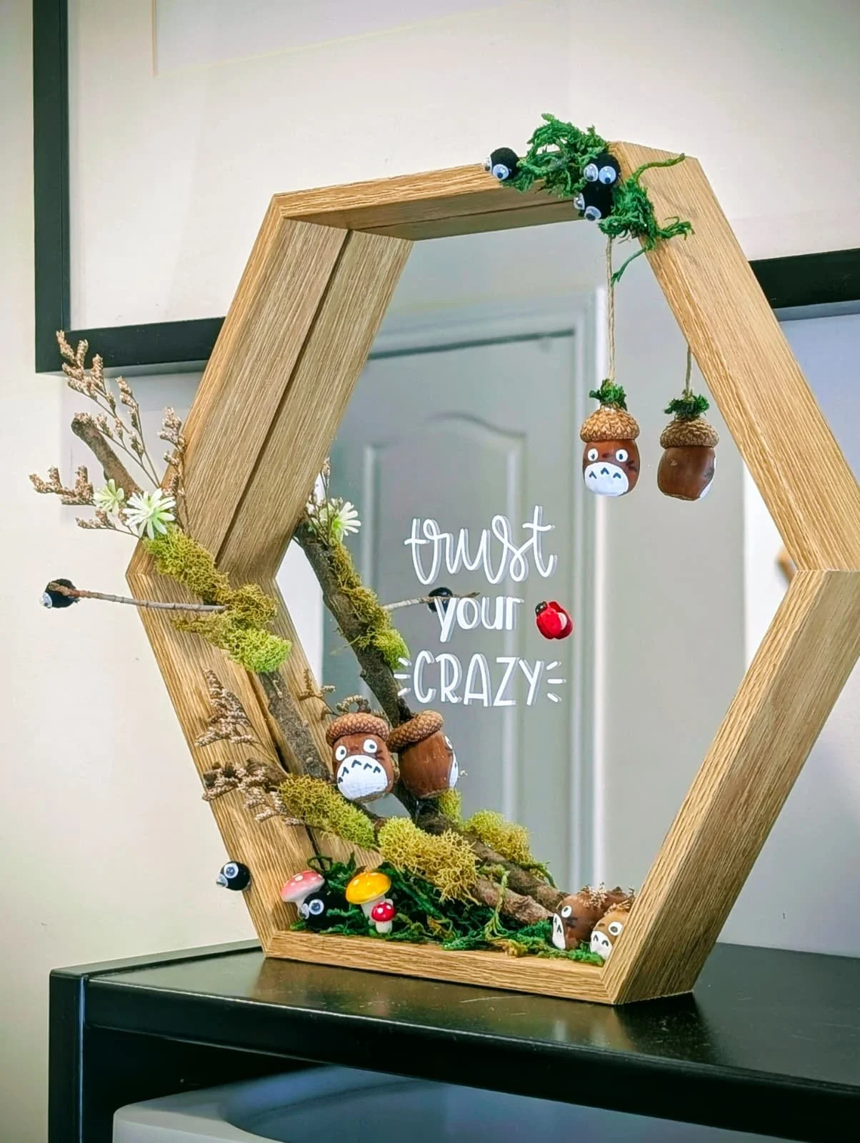 Decorative display with wooden hexagonal frame, faux foliage, small mushroom and acorn figurines with googly eyes, and a sign that reads "trust your CRAZY".