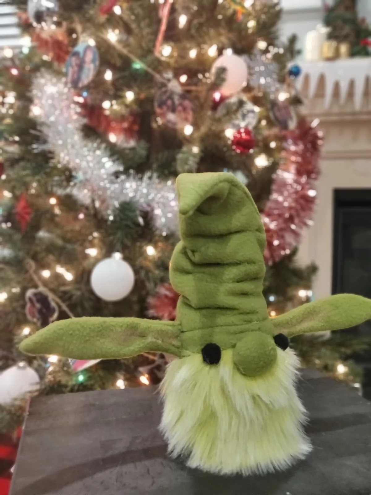 A green plush toy with a long body and large floppy arms, resembling a snake or dragon, sitting on a gray surface in front of a decorated Christmas tree with lights, ornaments, tinsel, and a blurred background.