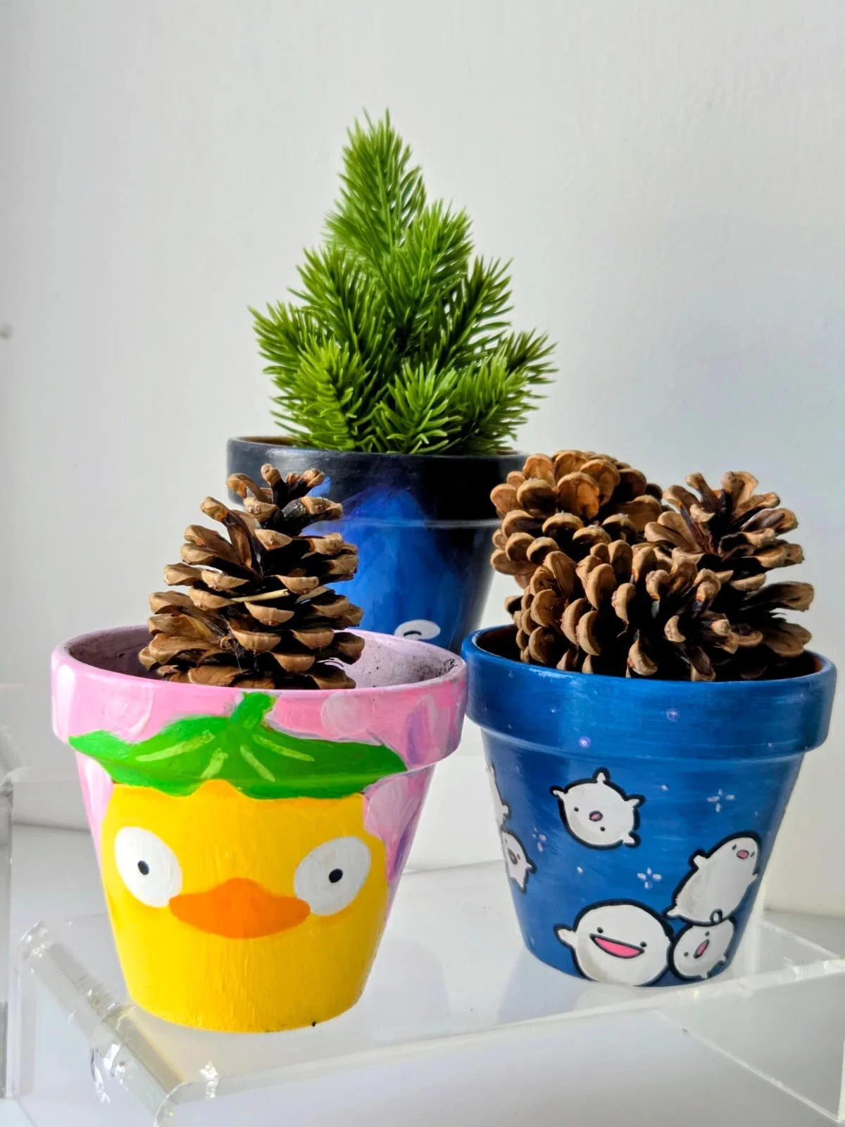 Three decorated plant pots with pinecones and a small artificial green pine in a dark blue pot, set on a white surface against a plain wall.