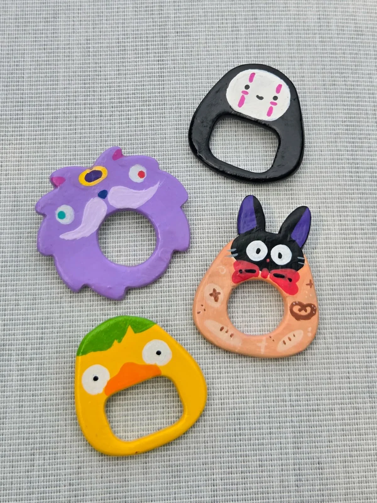 Colorful acrylic keychains on a textured gray surface, featuring a purple cat, a black and white creature with a face, a beige cat with a red bow, a yellow chick, and a black and white bunny.