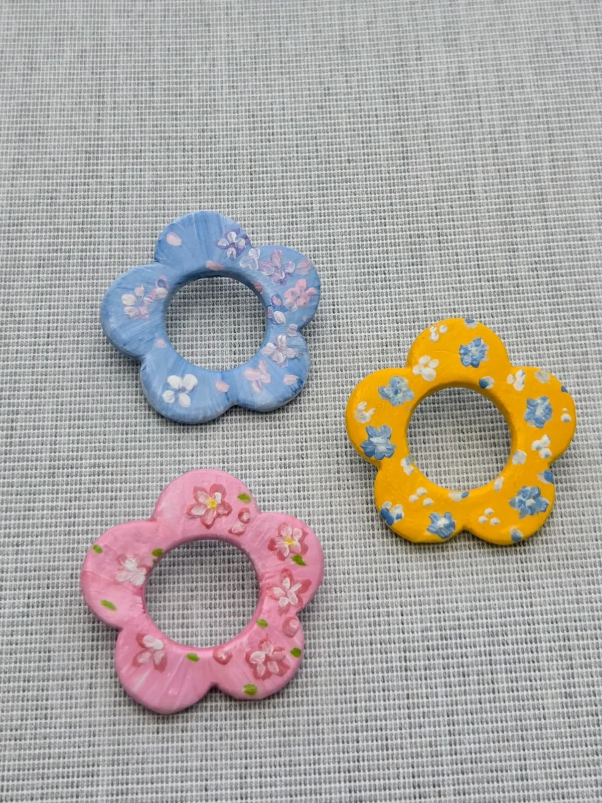 Three flower-shaped clips in pink, blue, and yellow with floral patterns on a gray textured surface.