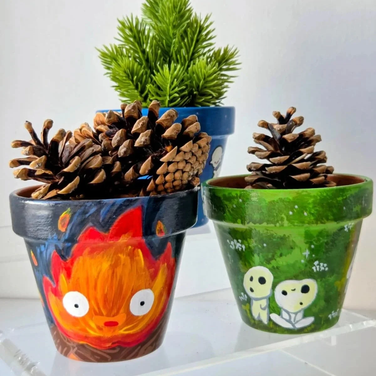 Decorative painted flower pots with pinecones and small green plants, one pot featuring a colorful fiery face and another with characters from the Peanuts comic strip.