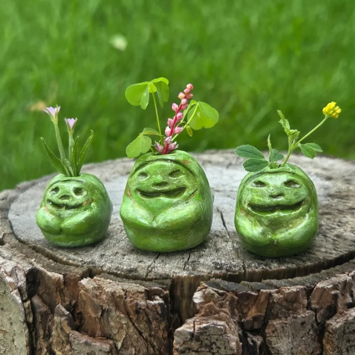Three carved green gourds with faces, each with flowers growing out of the top, placed on a tree stump with a grassy background.