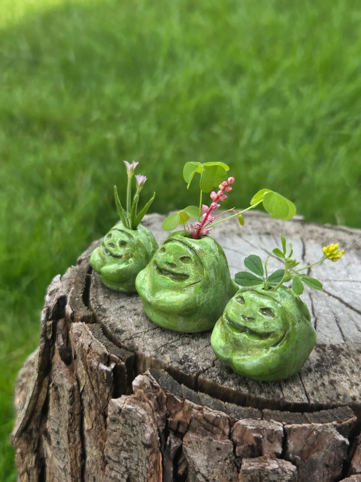 Three small green garden gnome sculptures with smiling faces, each with plants growing from the top of their heads, placed on a weathered tree stump, with a grassy background.