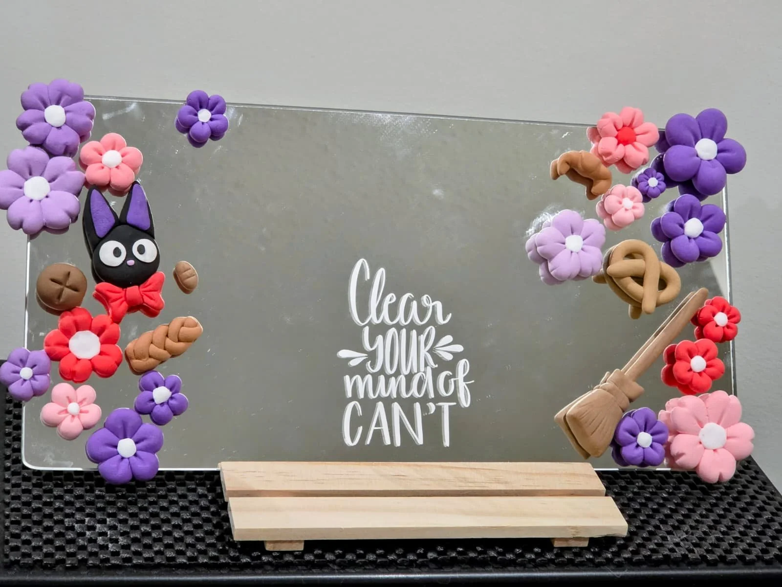 Decorative mirror with colorful clay flowers, a black cat figure with purple ears and a red bow, and a wooden stand, with a message that reads 'Clear your mug on, can't'.