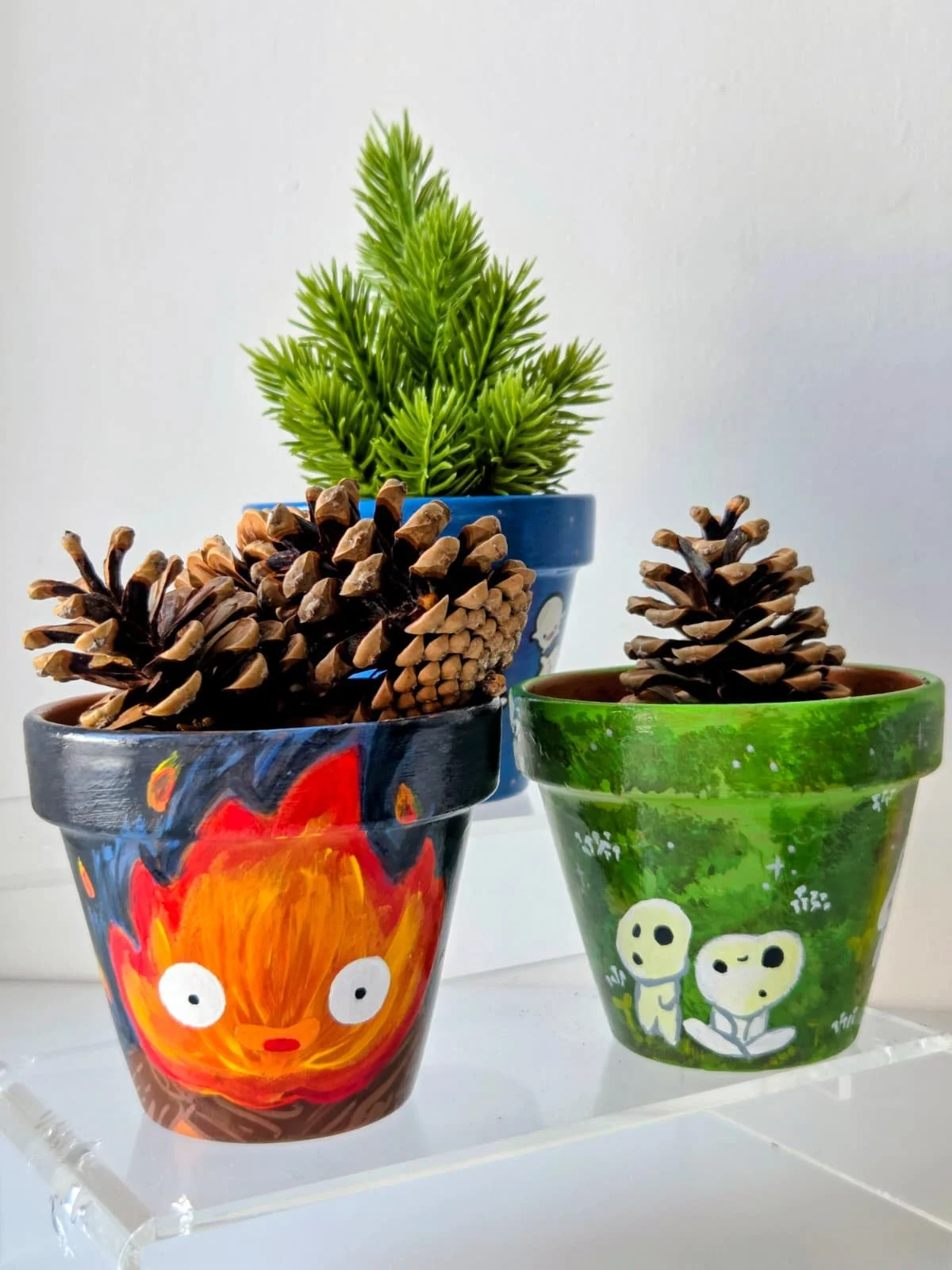 Three decorated flowerpots, each with pinecones, with the back pot containing a small green artificial pine tree. The front left pot has a hand-painted colorful design with a fiery face, and the front right pot has a green leafy pattern with cartoon-