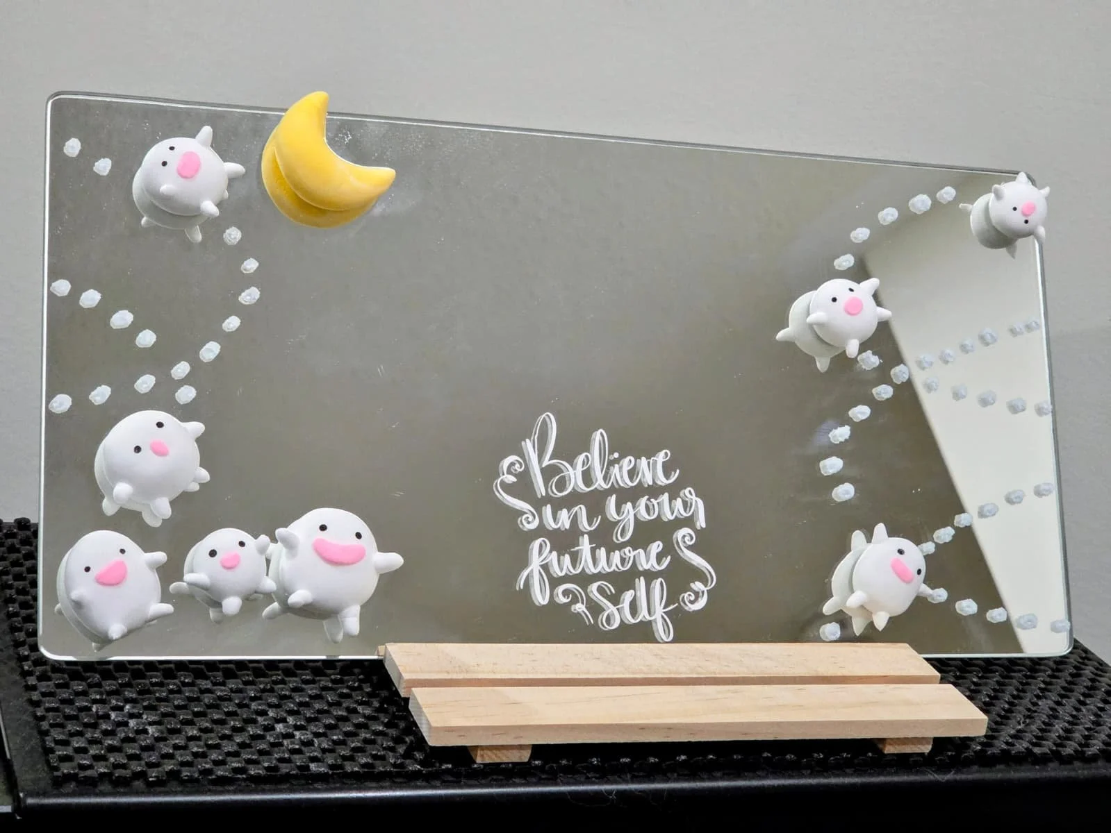 Decorative mirror with white bunny and yellow crescent moon magnets, featuring a motivational quote that says 'Believe in your future self.' The mirror is placed on a wooden stand on a textured black surface.
