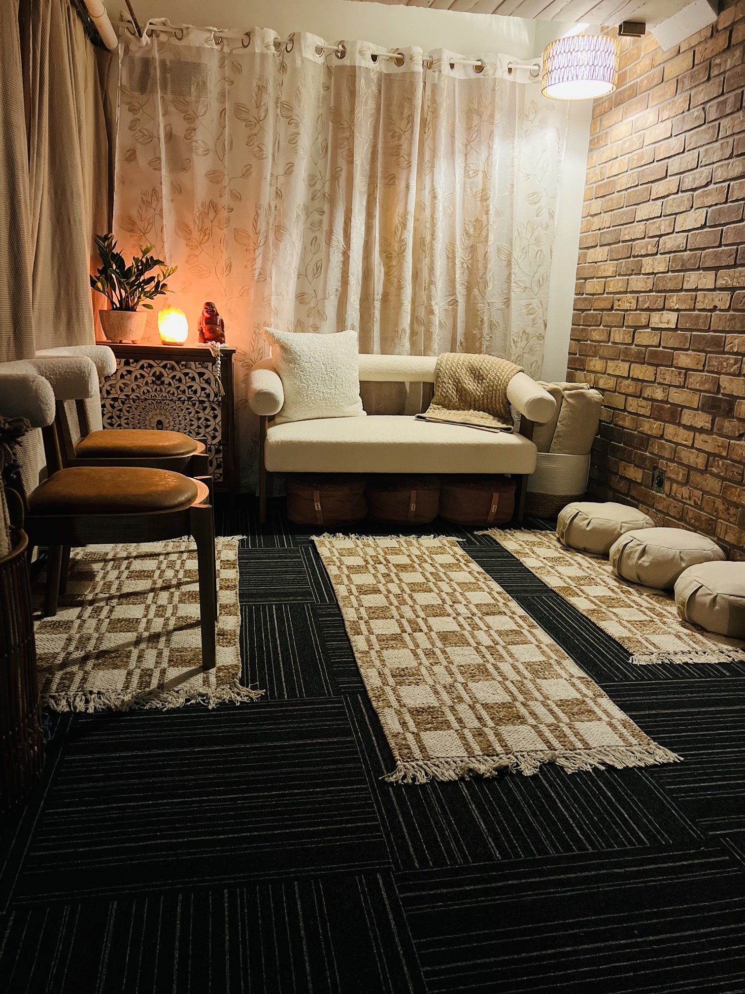 A cozy living room corner with a white sofa adorned with a textured pillow and a knitted blanket, positioned in front of a brick wall and cream curtains. A side table with a salt lamp, a salt lamp, and a small statue of a dog are visible. The room features a black and beige patterned rug and has a warm, inviting ambiance.