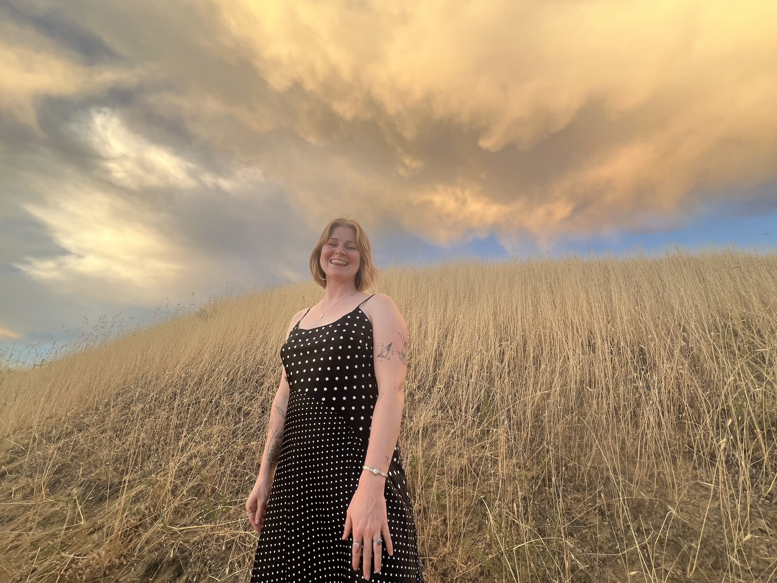A woman standing in a field of tall grass with a dramatic sky filled with large, yellow-orange clouds overhead, smiling.
