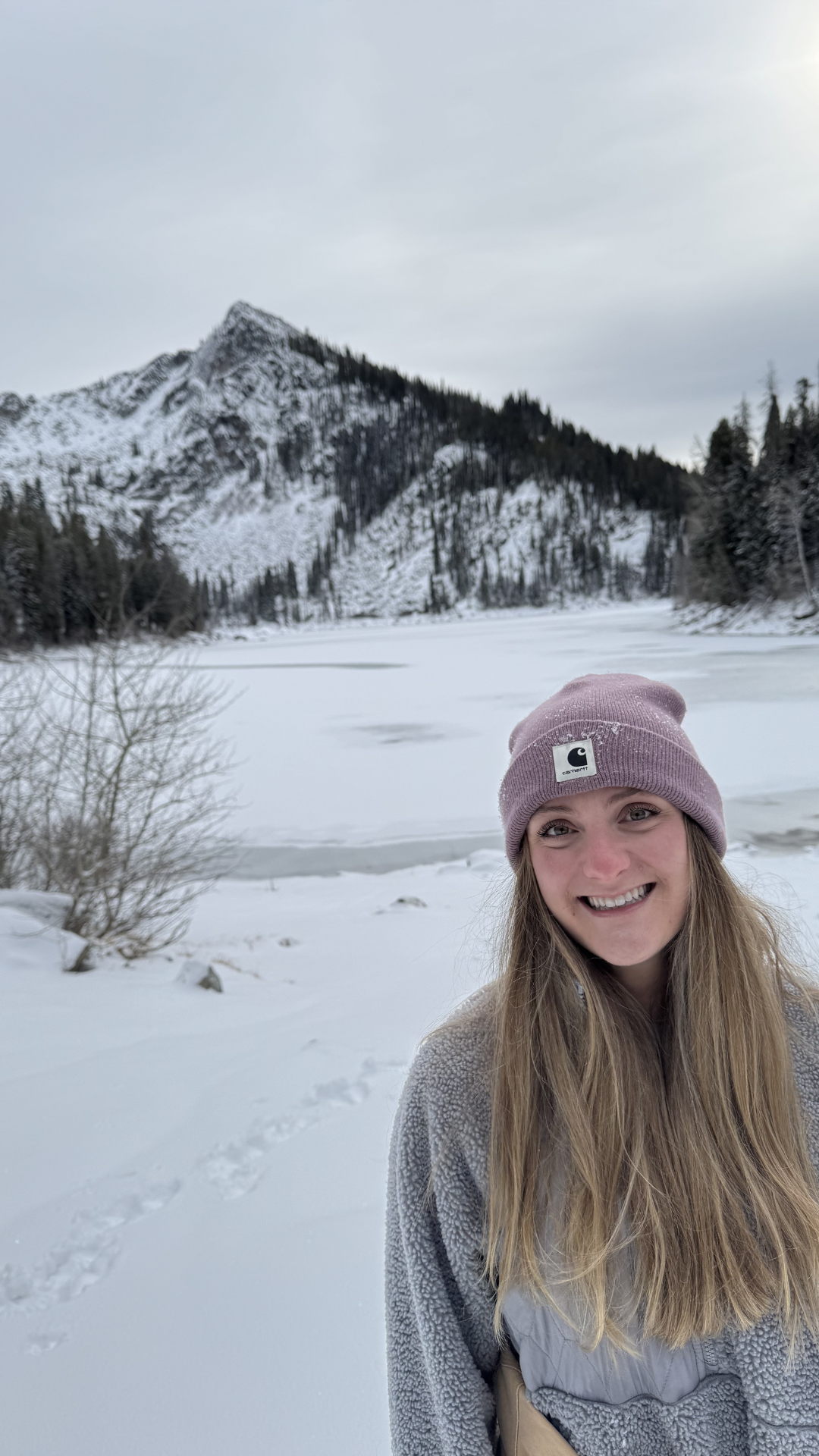 A young woman smiling outdoors in a snowy, mountainous landscape, wearing a pink beanie and a gray fleece jacket.