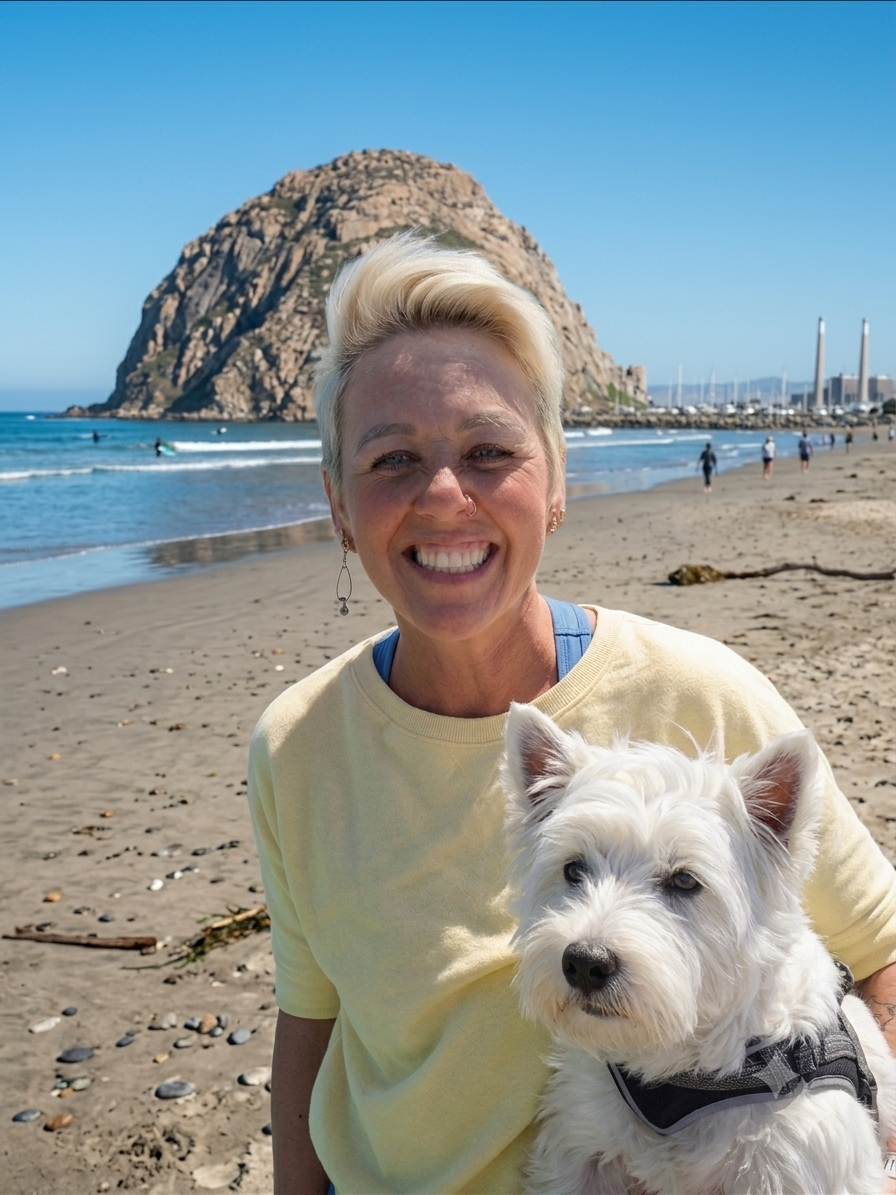 A woman with short blond hair smiling at the camera, holding a small white dog on a beach with a large rocky hill and a marina in the background.