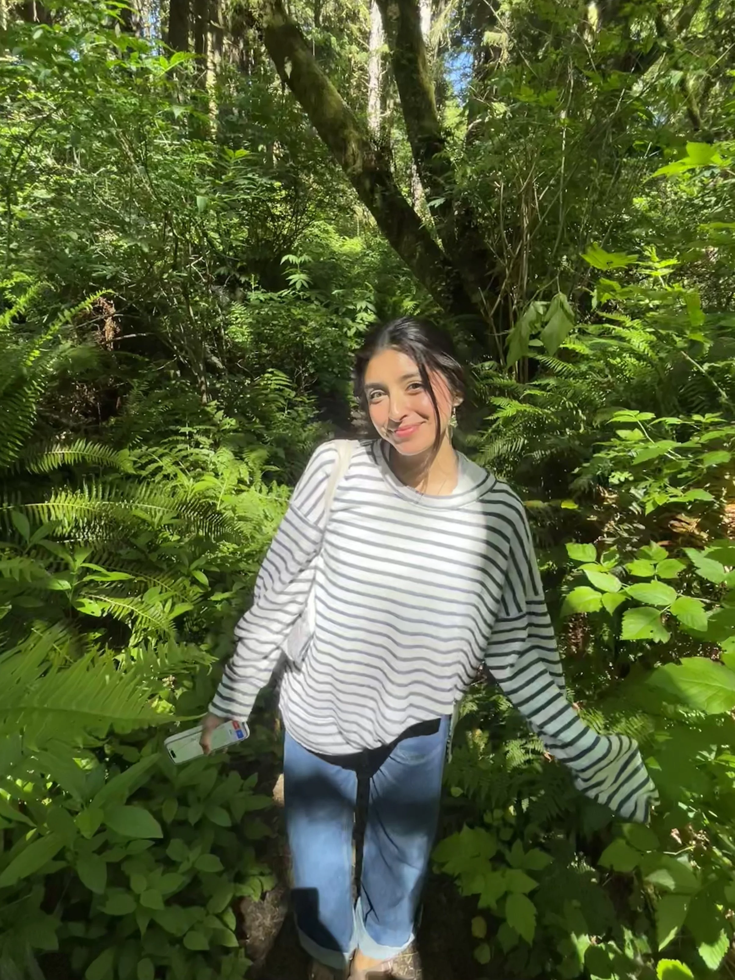 A smiling woman with dark hair in a striped long-sleeve shirt and blue jeans standing among lush green foliage and trees in a forest or jungle setting, holding a smartphone in her right hand.