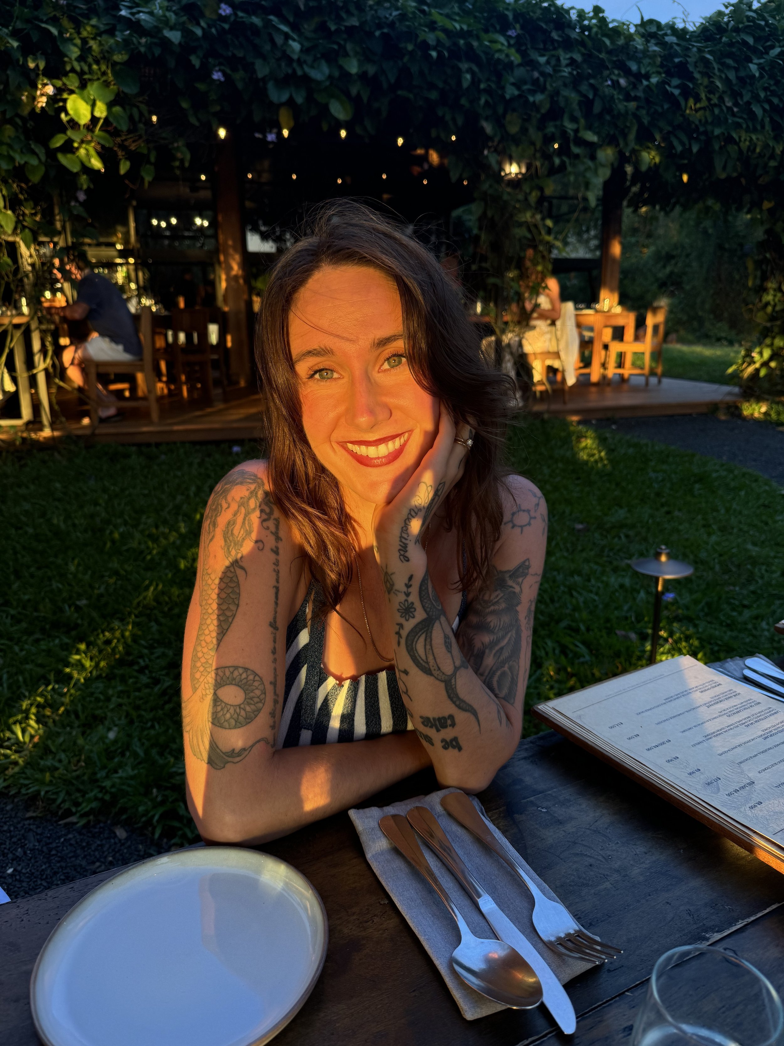 A smiling woman with tattoos on her arms sitting at an outdoor restaurant table during sunset. The table has empty plates and utensils. In the background, there are other diners and a green outdoor setting.