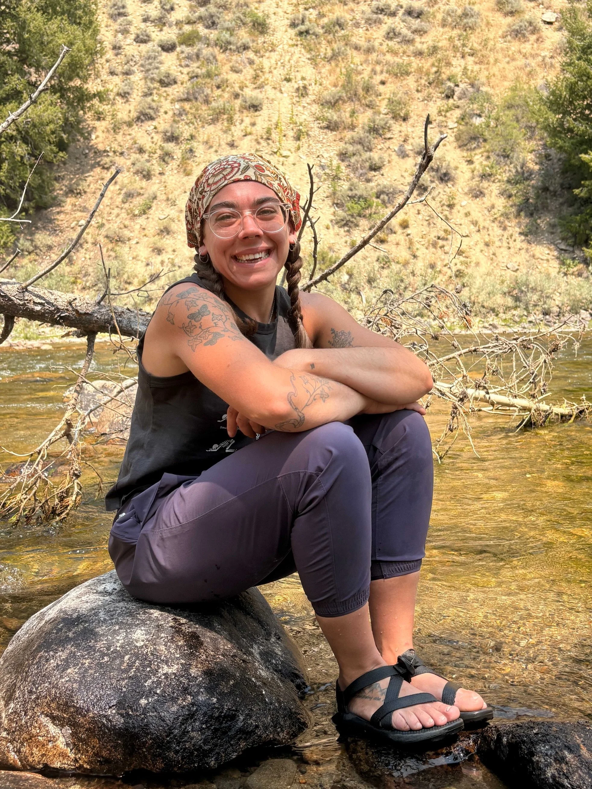 A smiling woman with tattoos, wearing glasses, a patterned headscarf, a black sleeveless shirt, and dark pants, sitting on a rock in a shallow creek with a yellow hillside and green bushes in the background.