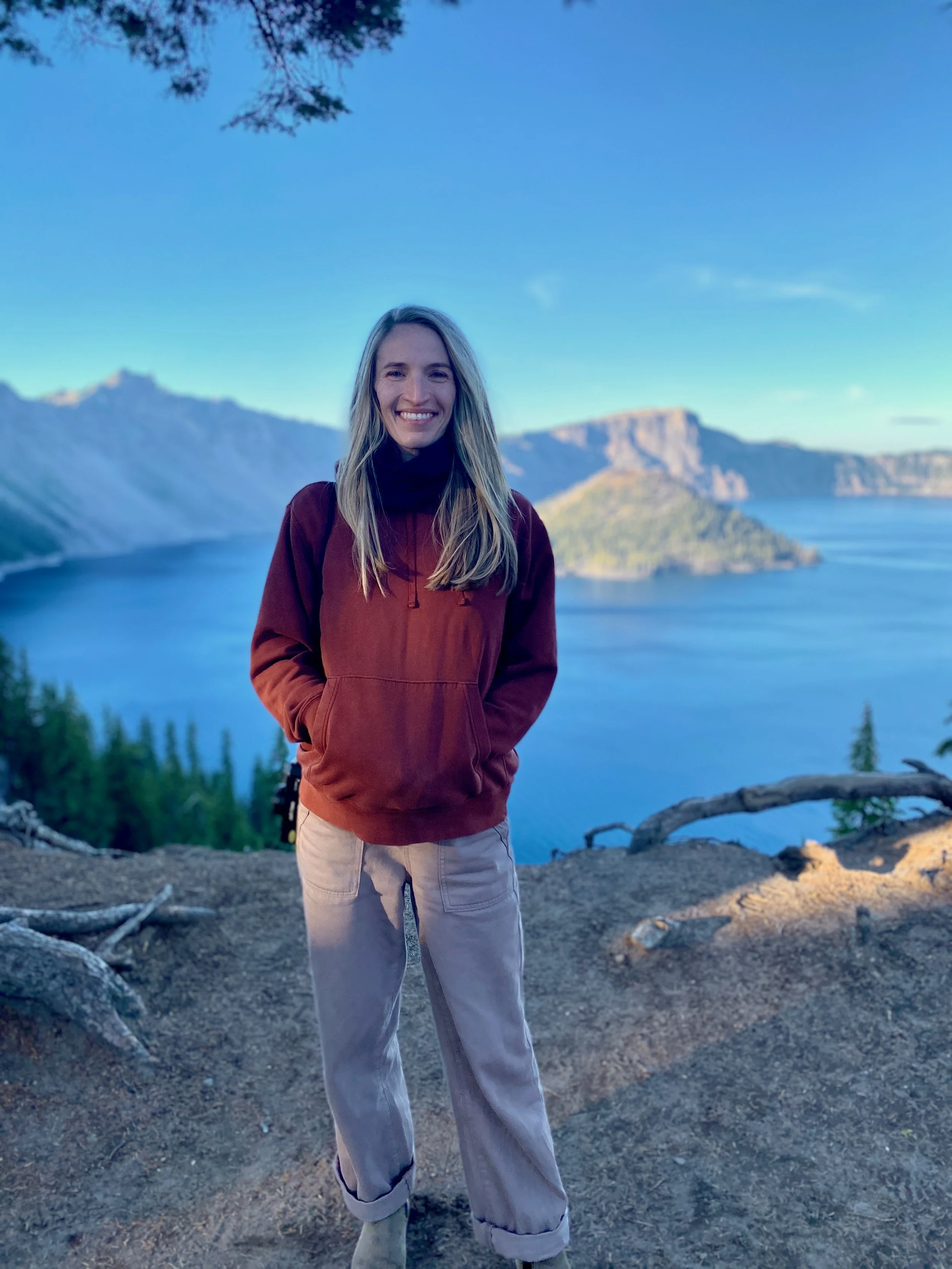 A young woman with long blonde hair smiling outdoors, standing on rocky ground with a scenic view of a lake and mountains in the background under a clear blue sky.