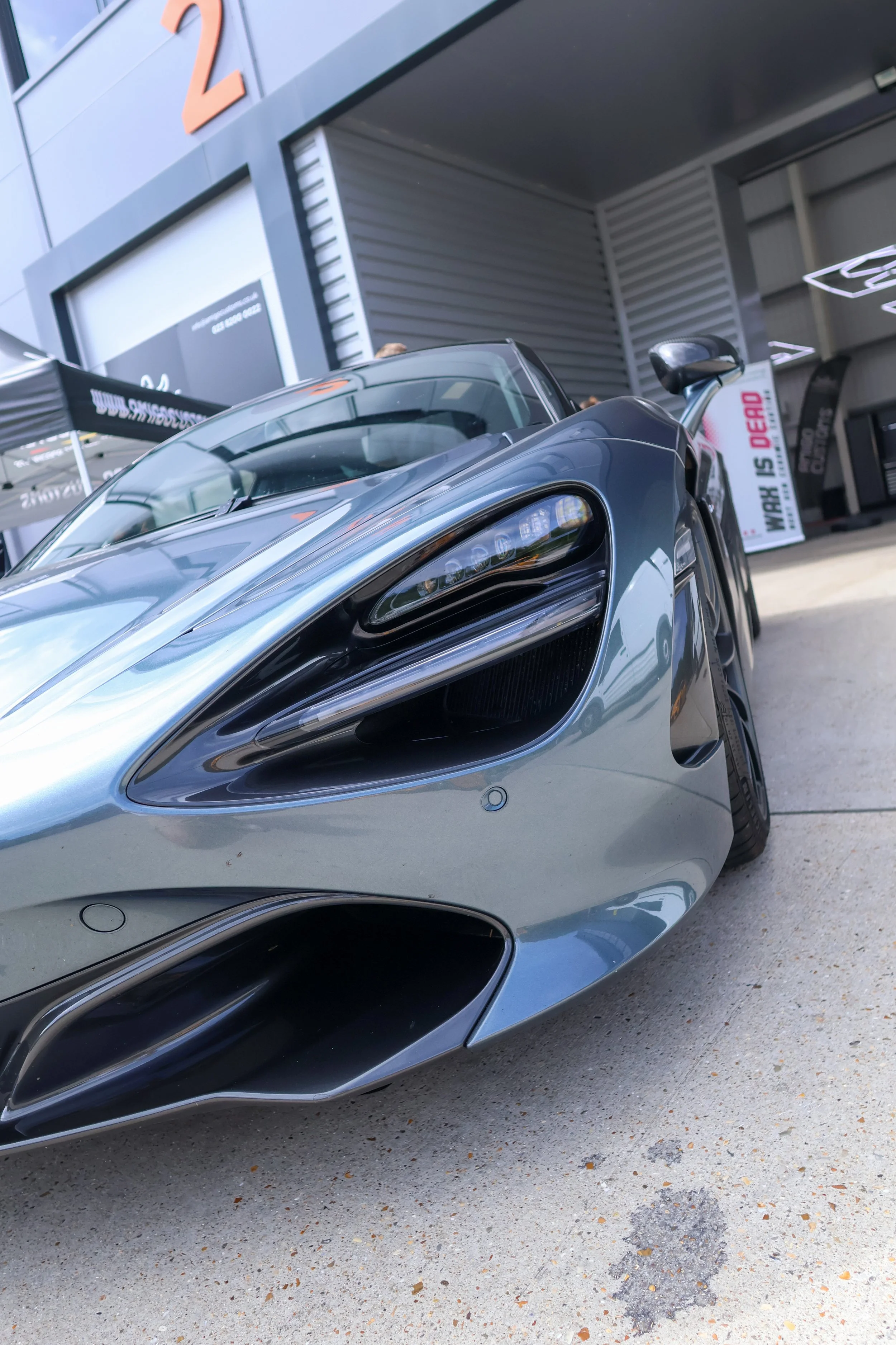 Close-up of a sleek, silver sports car with aerodynamic design and advanced headlights, parked outside a modern building with a large orange number '2' on the wall.
