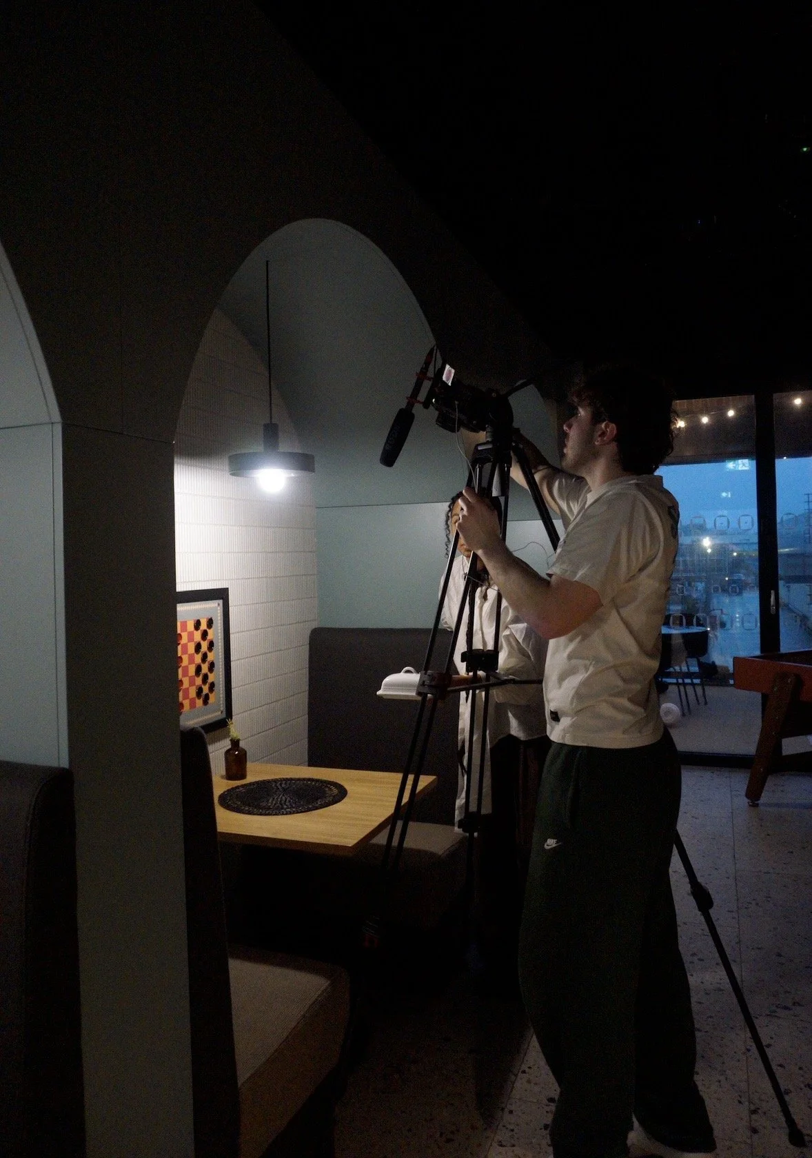 Person filming in dimly lit room with a tripod near a table and window, at night.