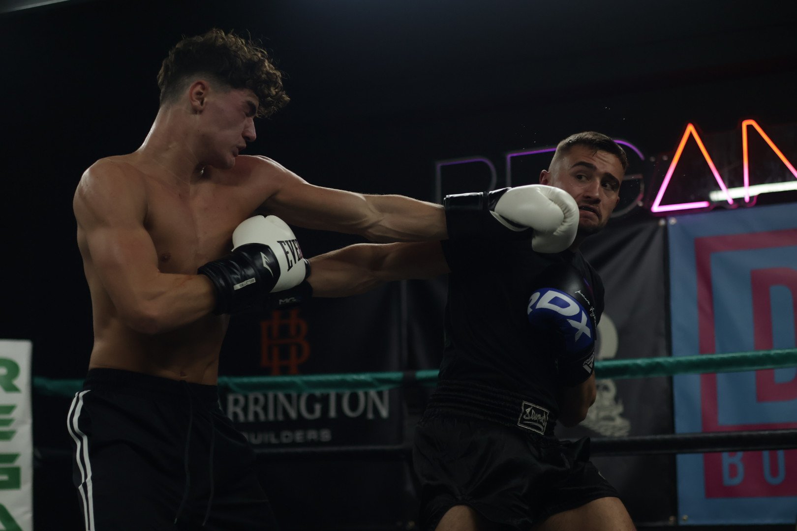 Two male boxers in a boxing ring engaged in a fight. The boxer on the left, shirtless, is landing a punch on the opponent on the right, who is wearing a black shirt. The boxer on the right has a distressed expression as the punch connects.