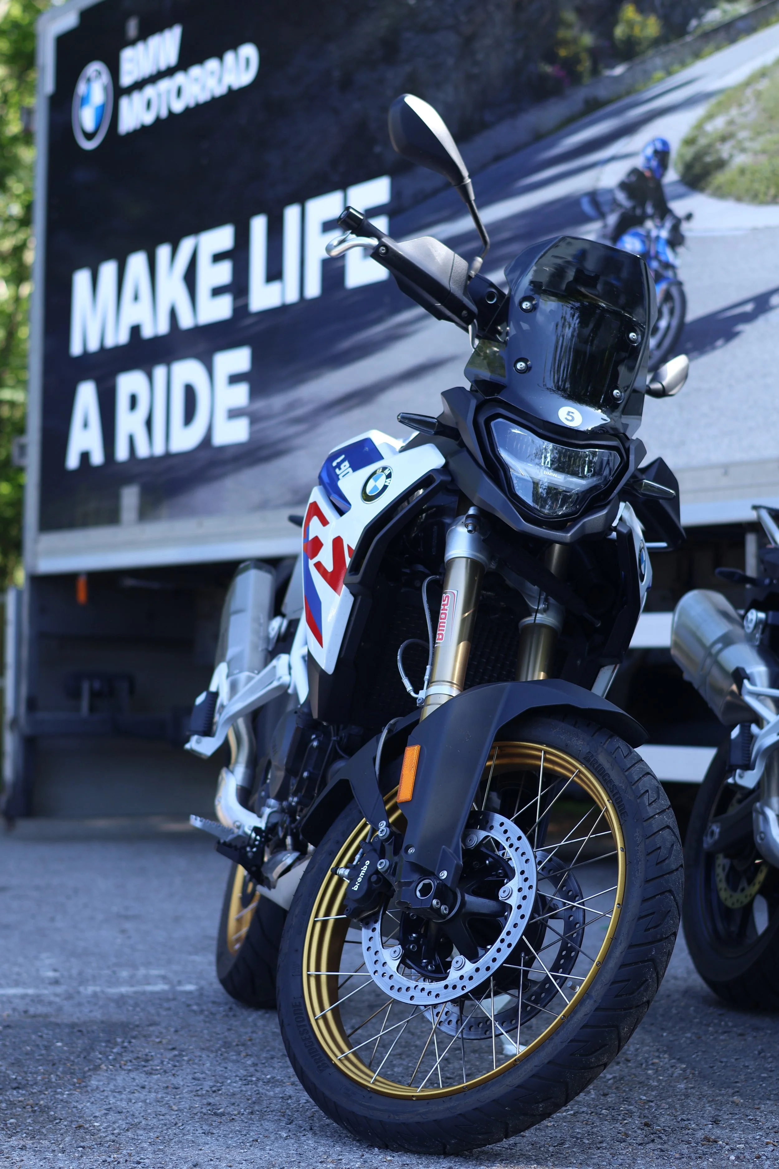 Close-up view of a BMW motorcycle with gold rims, black body, and windshield, parked in front of a large billboard with motorcycle riders and the text 'BMW MOTORRAD MAKE LIFE A RIDE.'