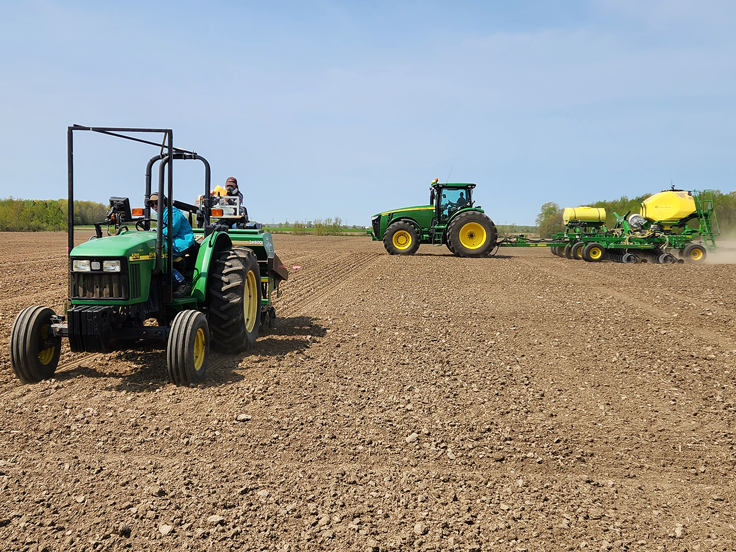 Farmers planting grain tractors in a large field in Conn Ontario, with one tractor pulling a planter or spray equipment, all under a clear sky.