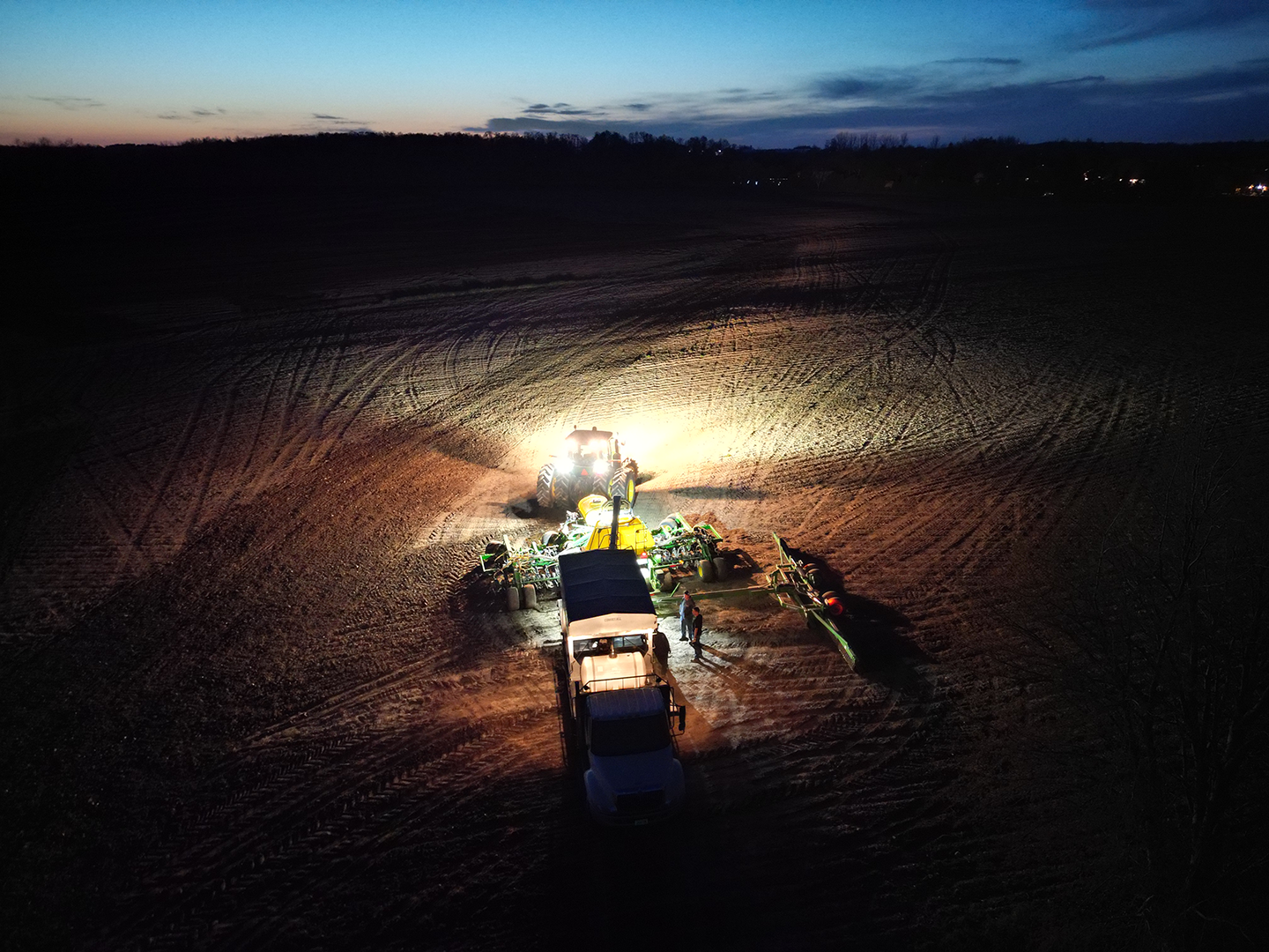 A large tractor and farming equipment on a dirt field at dusk, with a truck and two people nearby illuminated by bright lights.