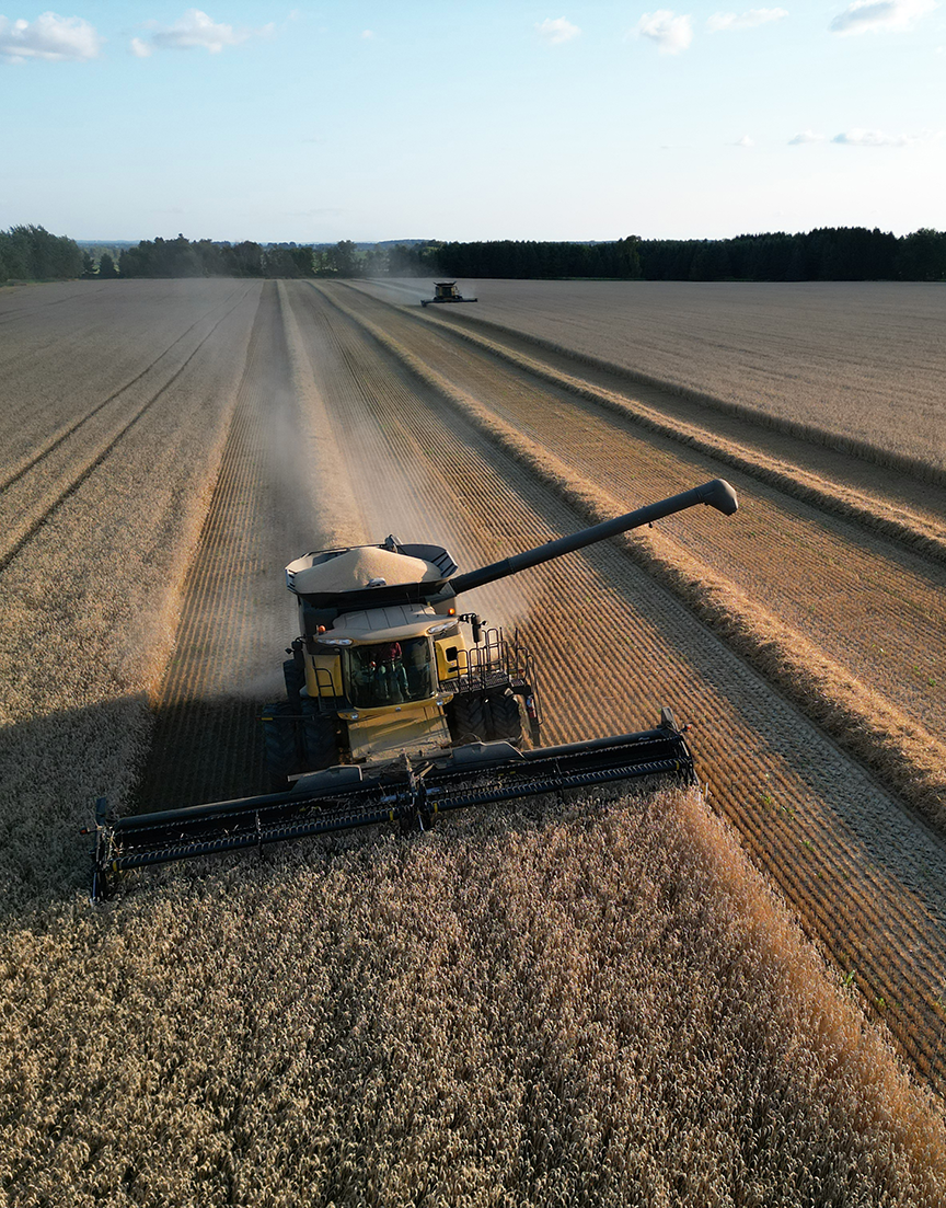 A combine harvester working in a large wheat field during daytime, with another harvester in the distance, under a partly cloudy sky.