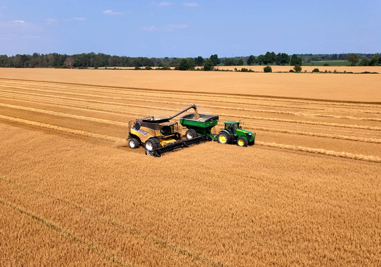 A New Holland combine offloading into a grain wagon being pulled by a John Deere tractor working in a wheat field, during daytime under clear blue skies.
